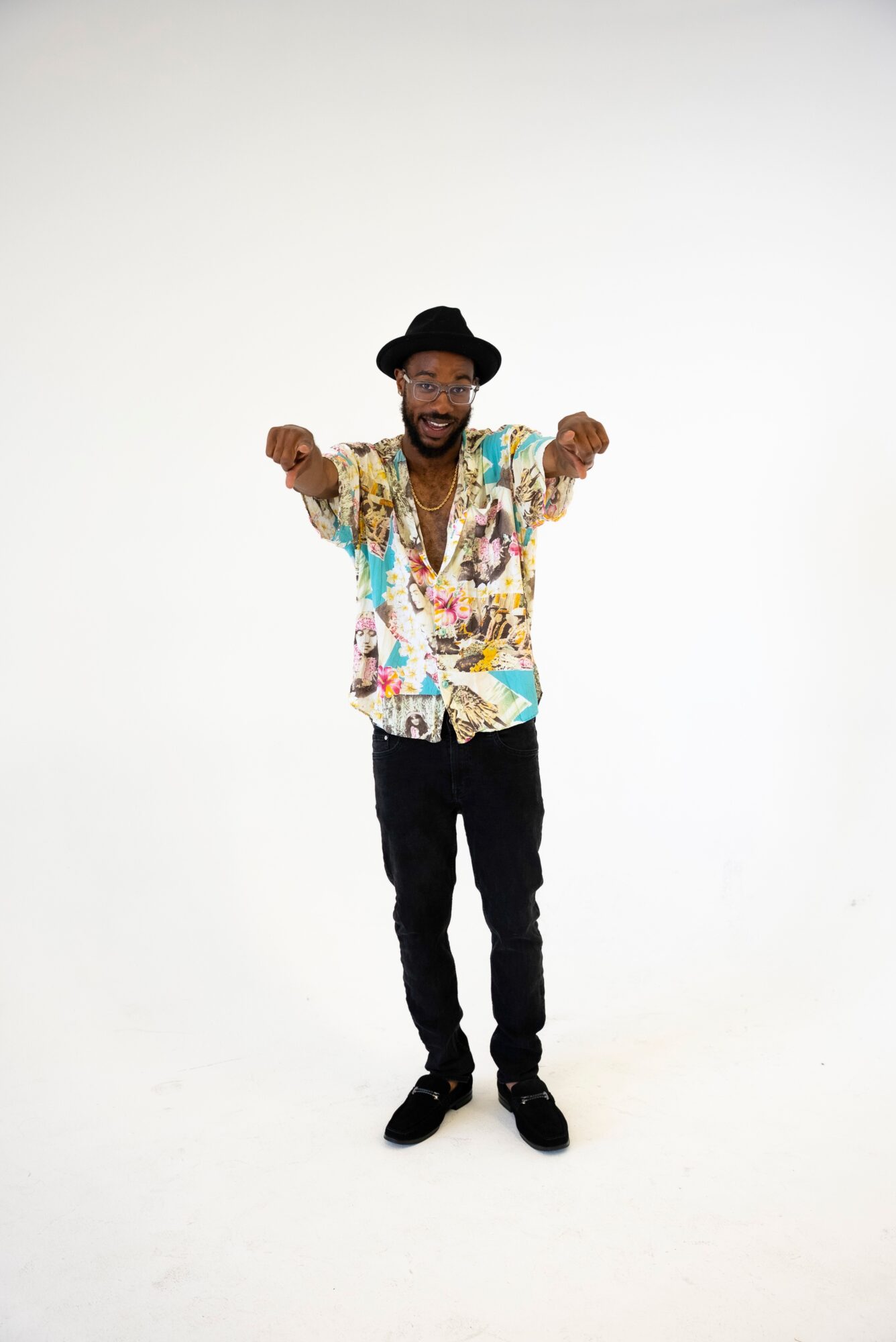 Person smiling and pointing forward, wearing a colorful shirt, black pants, and a black hat, standing against a white background.