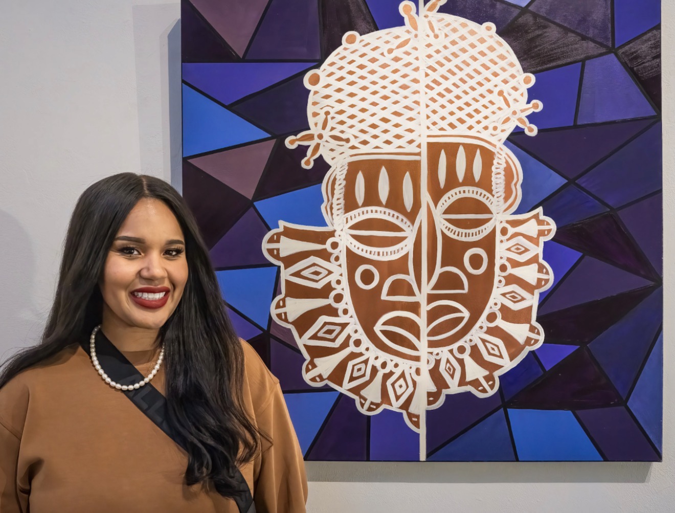 Woman with long dark hair, wearing a brown top and pearl necklace, standing in front of colorful geometric art with a face design.