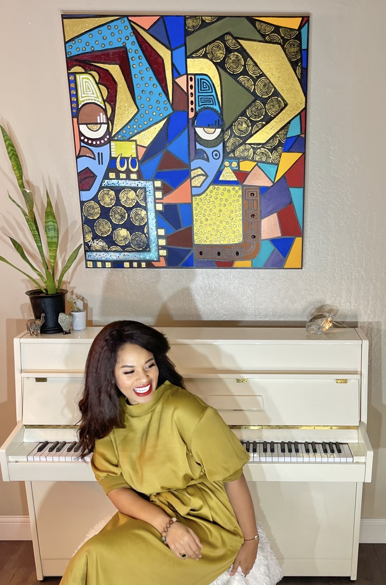 Woman in yellow dress sitting on a white piano, colorful abstract painting on wall behind her, potted plant on top of piano.