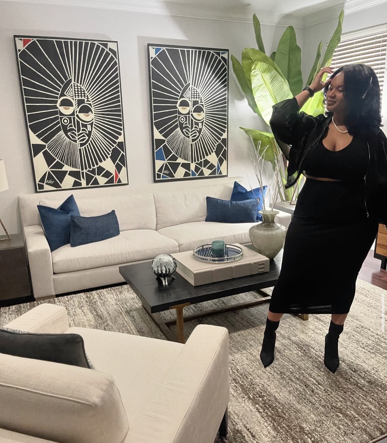 Woman in black dress and boots standing in living room with sofa, blue pillows, artwork, and large plant, smiling and touching her hair.