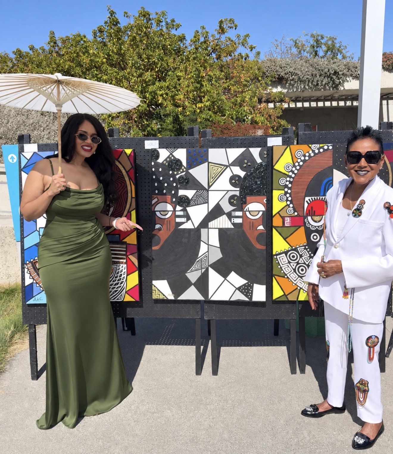 Two women stand outdoors near colorful abstract art panels, one in a green dress and the other in a white suit, smiling.