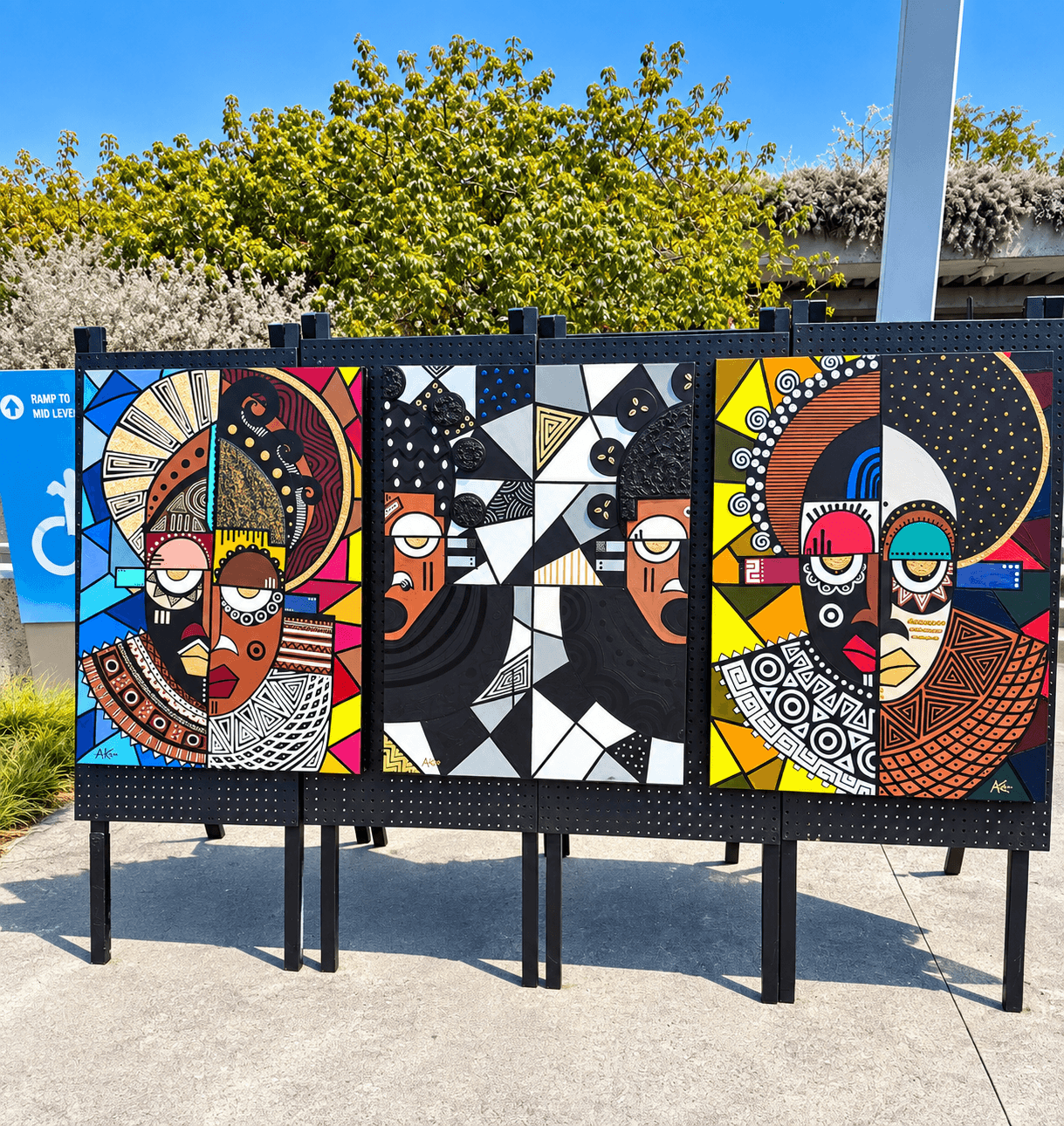 Three abstract portraits with geometric patterns and vibrant colors on display outdoors, with trees and a blue sky in background.