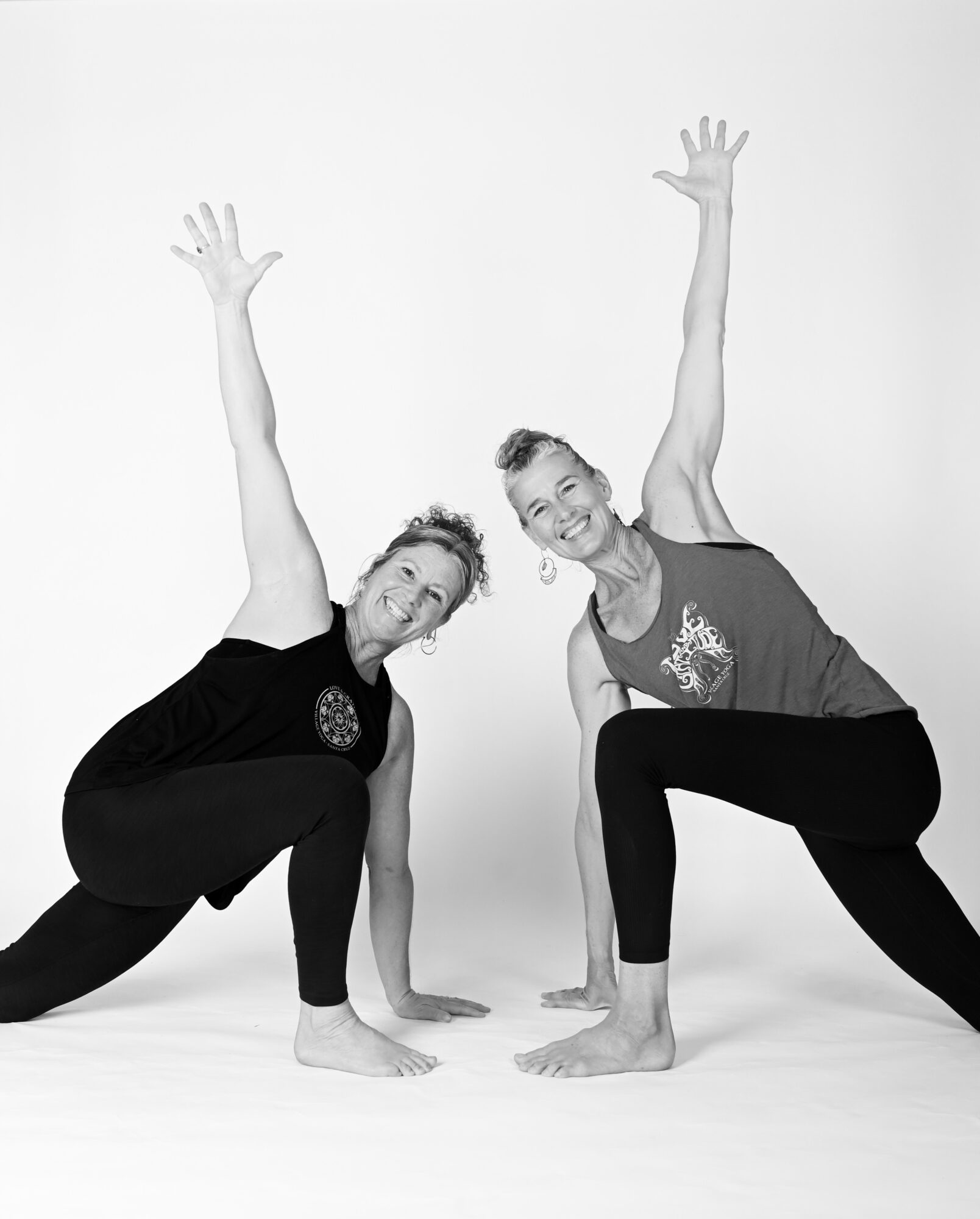 Two women in yoga poses with one arm raised and one hand on the ground, smiling, against a plain background.