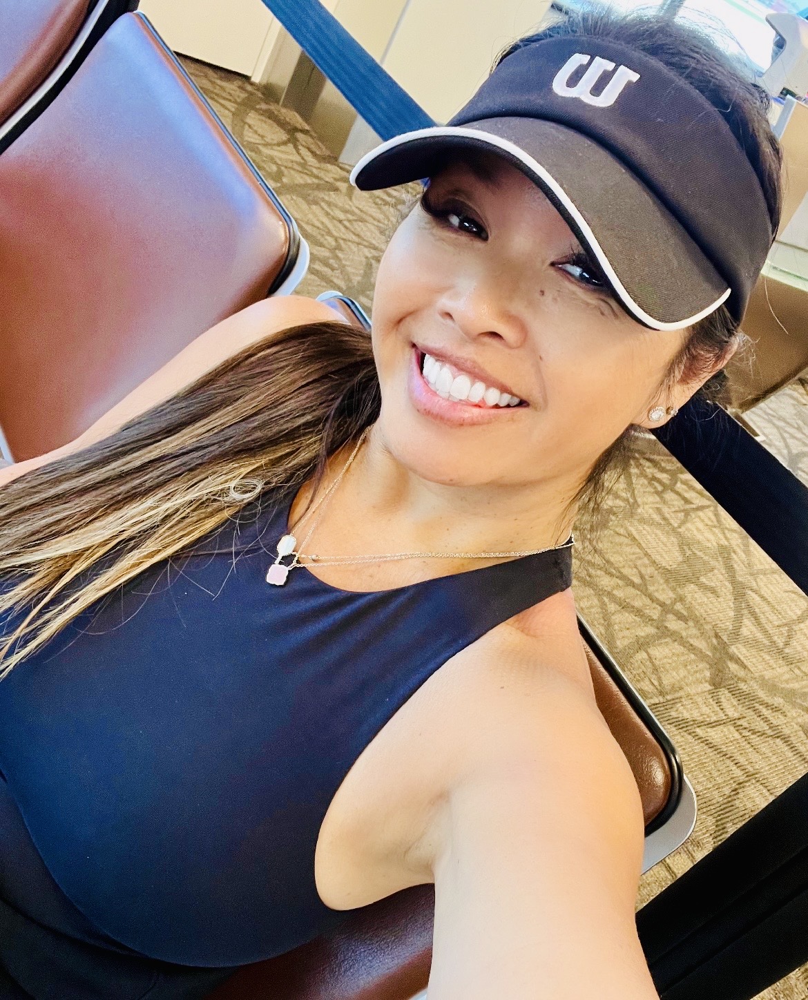 Smiling woman wearing a black cap and sleeveless top, sitting indoors, taking a selfie.