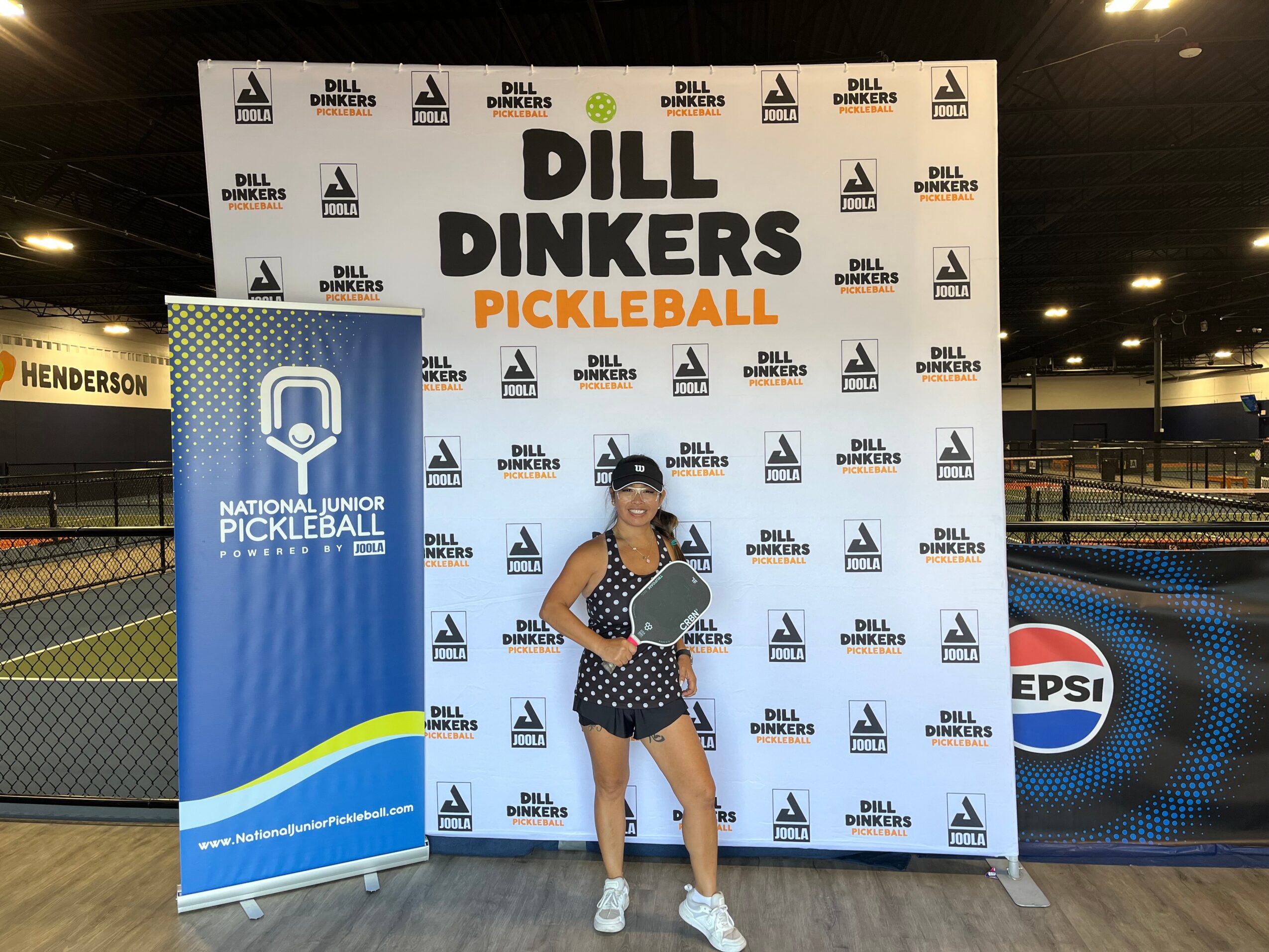 Person standing in front of a DILL DINKERS PICKLEBALL backdrop, wearing a mask, sunglasses, and casual clothing, holding a paddle.