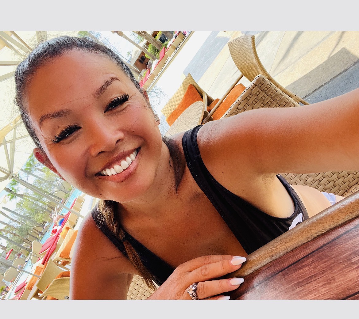 Smiling woman with dark hair in a ponytail, wearing a black sleeveless top, sitting outdoors at a table.