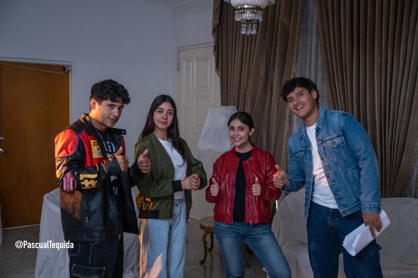Four young people standing indoors, smiling, with curtains and a chandelier in the background.