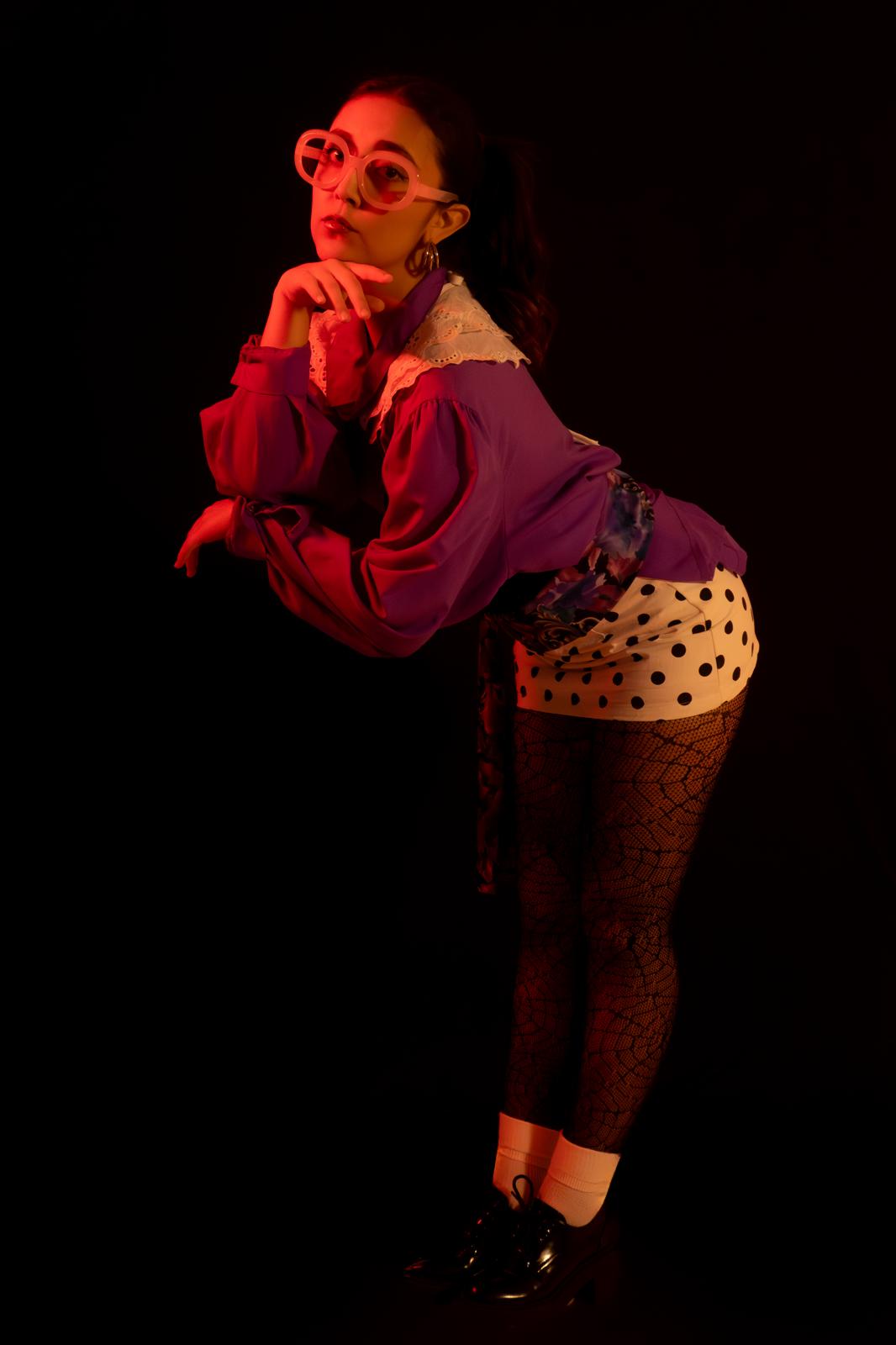 Person with glasses and colorful clothing poses with hand near face against dark background.