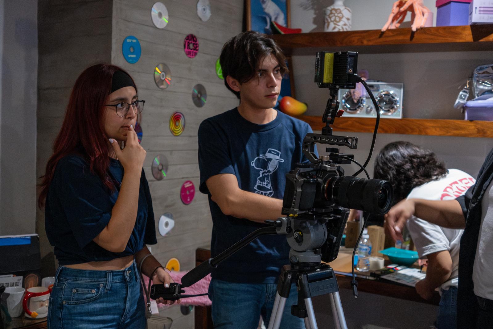Two young people operate a camera in a room with a wall decorated with colorful discs and shelves with objects.