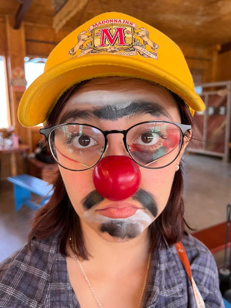 Person with clown makeup, red nose, glasses, yellow cap, indoors, serious expression.