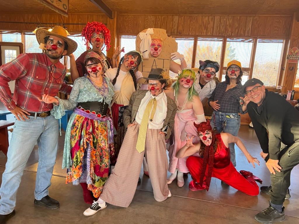 Group of people dressed as clowns and characters, posing indoors with wooden walls and windows in the background.