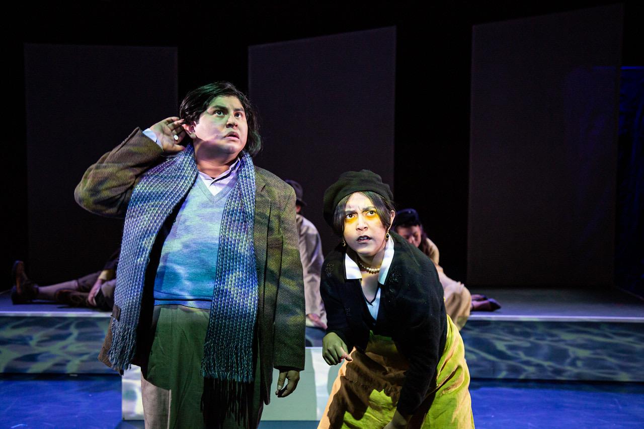 Two people on stage, one with a scarf, the other with glasses, both looking upward, with a dark background.