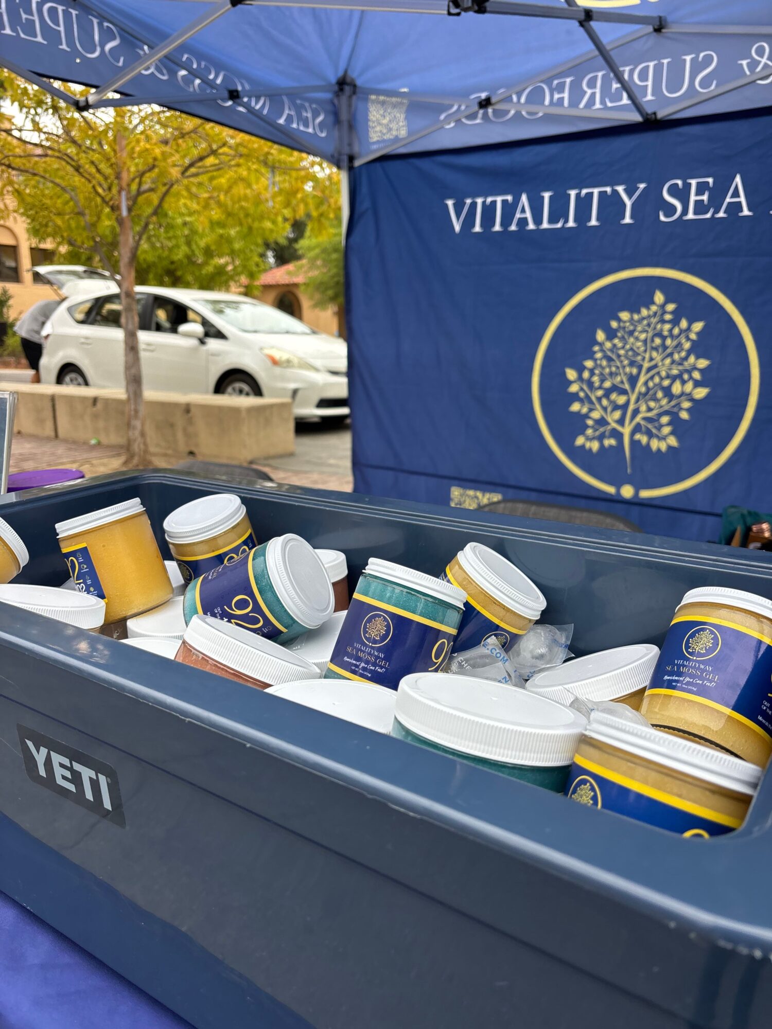 Open cooler with various supplement bottles and containers, outdoor parking lot with trees and cars in background.
