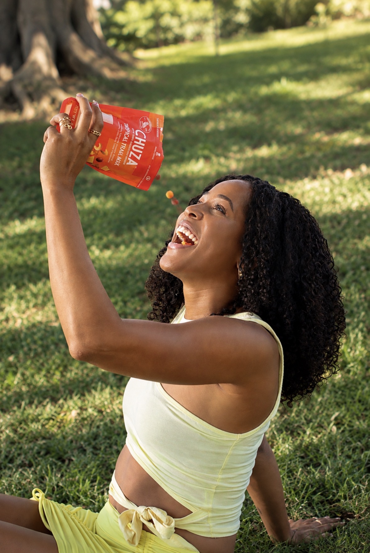 Woman with curly hair sitting on grass, pouring orange drink from a bottle, smiling, outdoors in sunlight.
