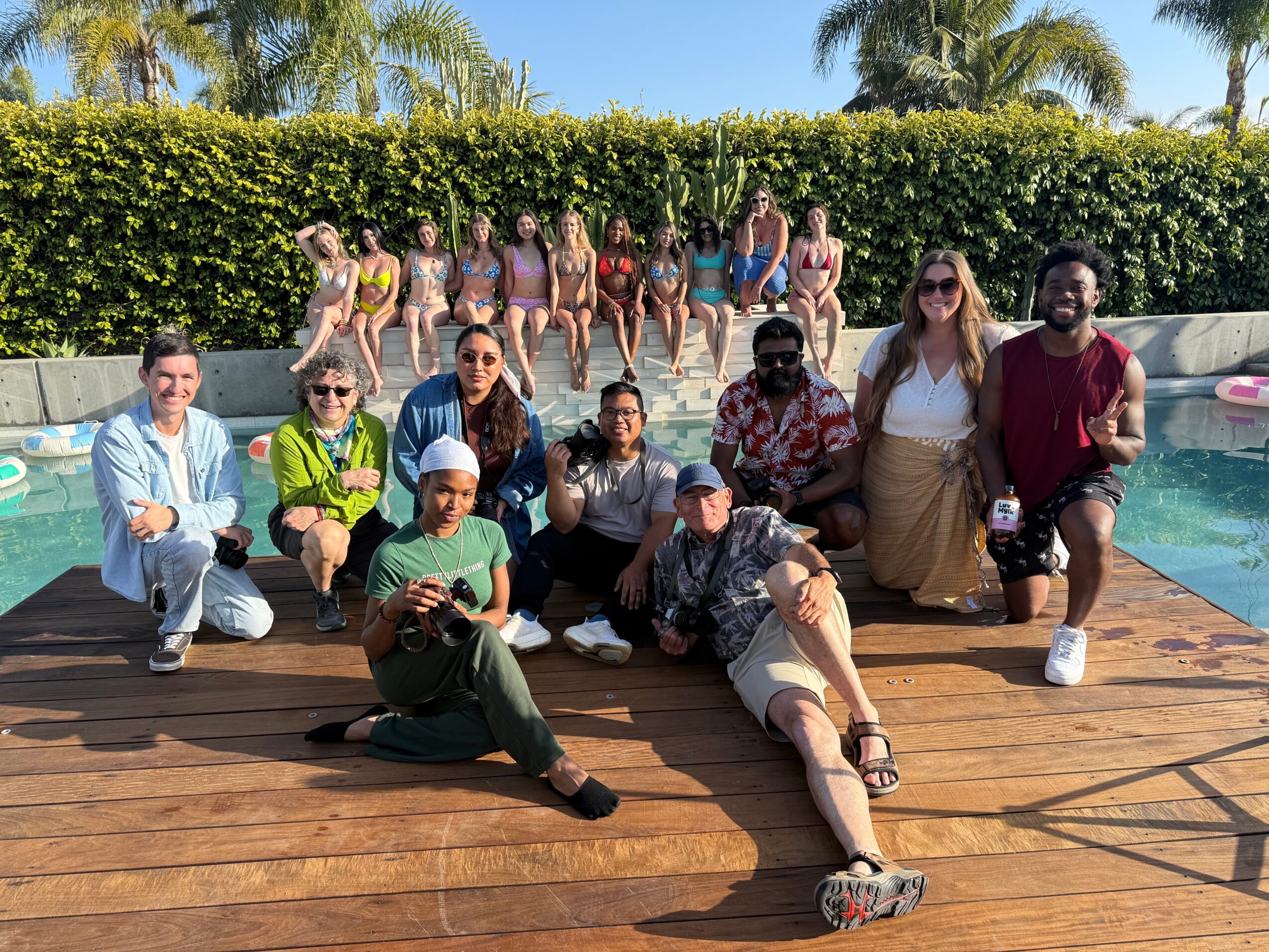 Group of people posing on a wooden deck near a pool with a hedge and palm trees in the background.