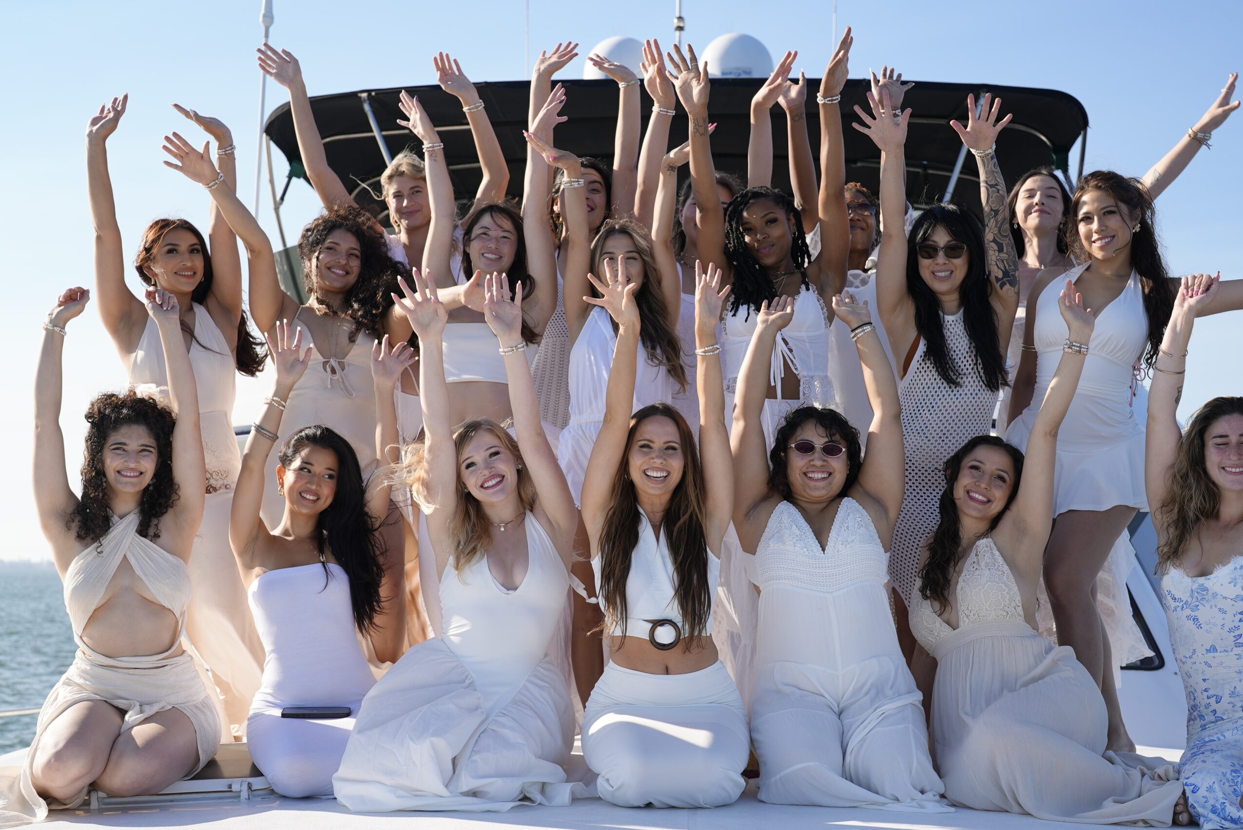 Group of women in white dresses raising hands outdoors on a boat deck, smiling, sunny day.