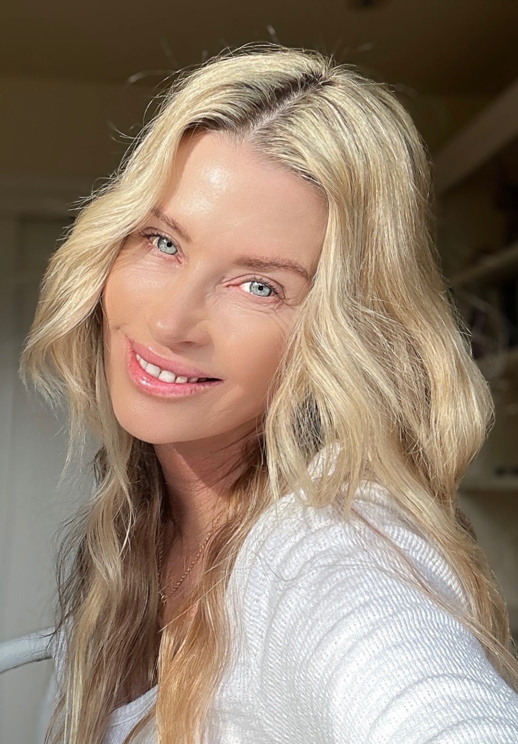 Woman with long wavy blonde hair smiling, wearing a white top, indoors with warm lighting.