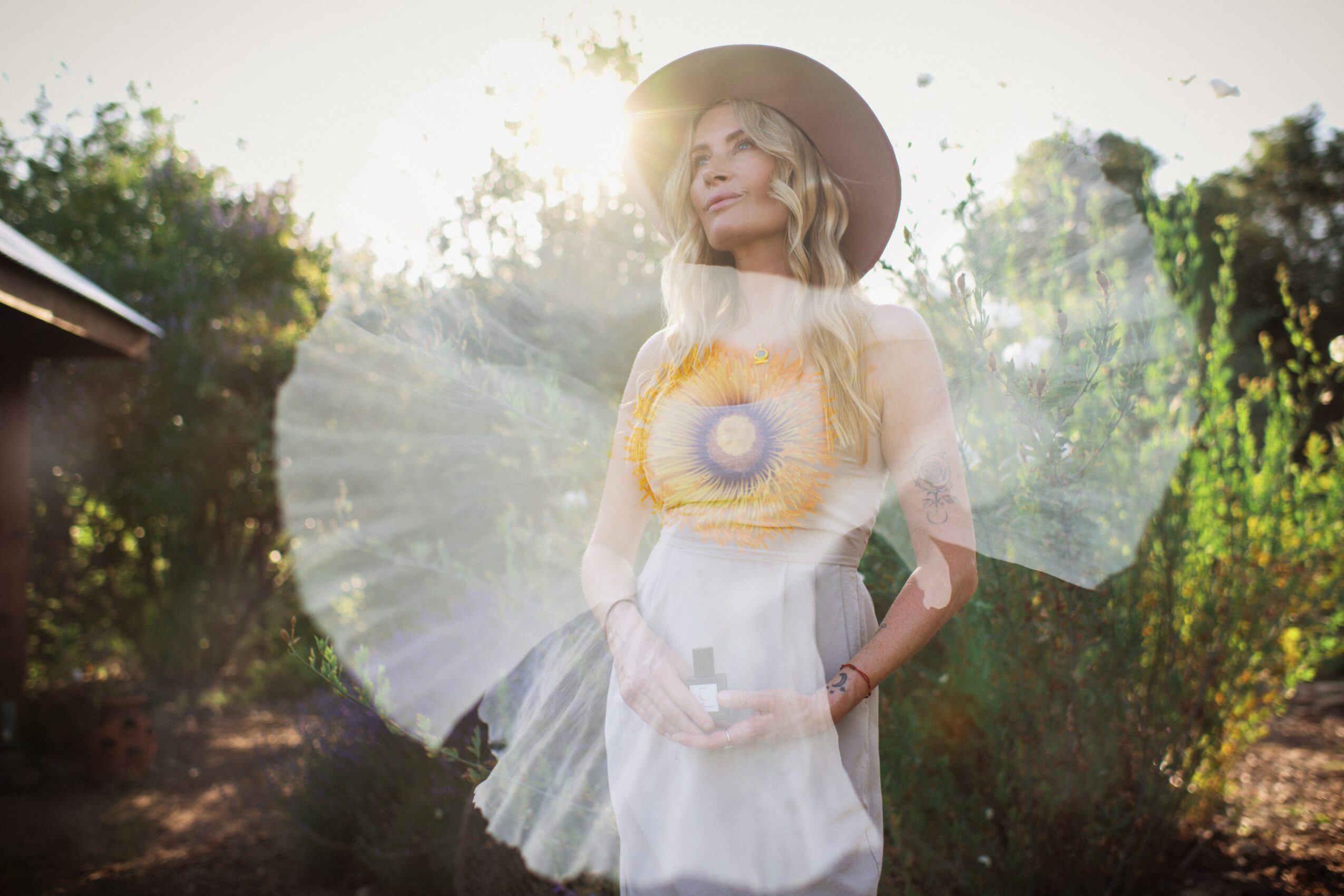 Woman with long blonde hair wearing a hat and a yellow top, standing outdoors with sunlight behind her.
