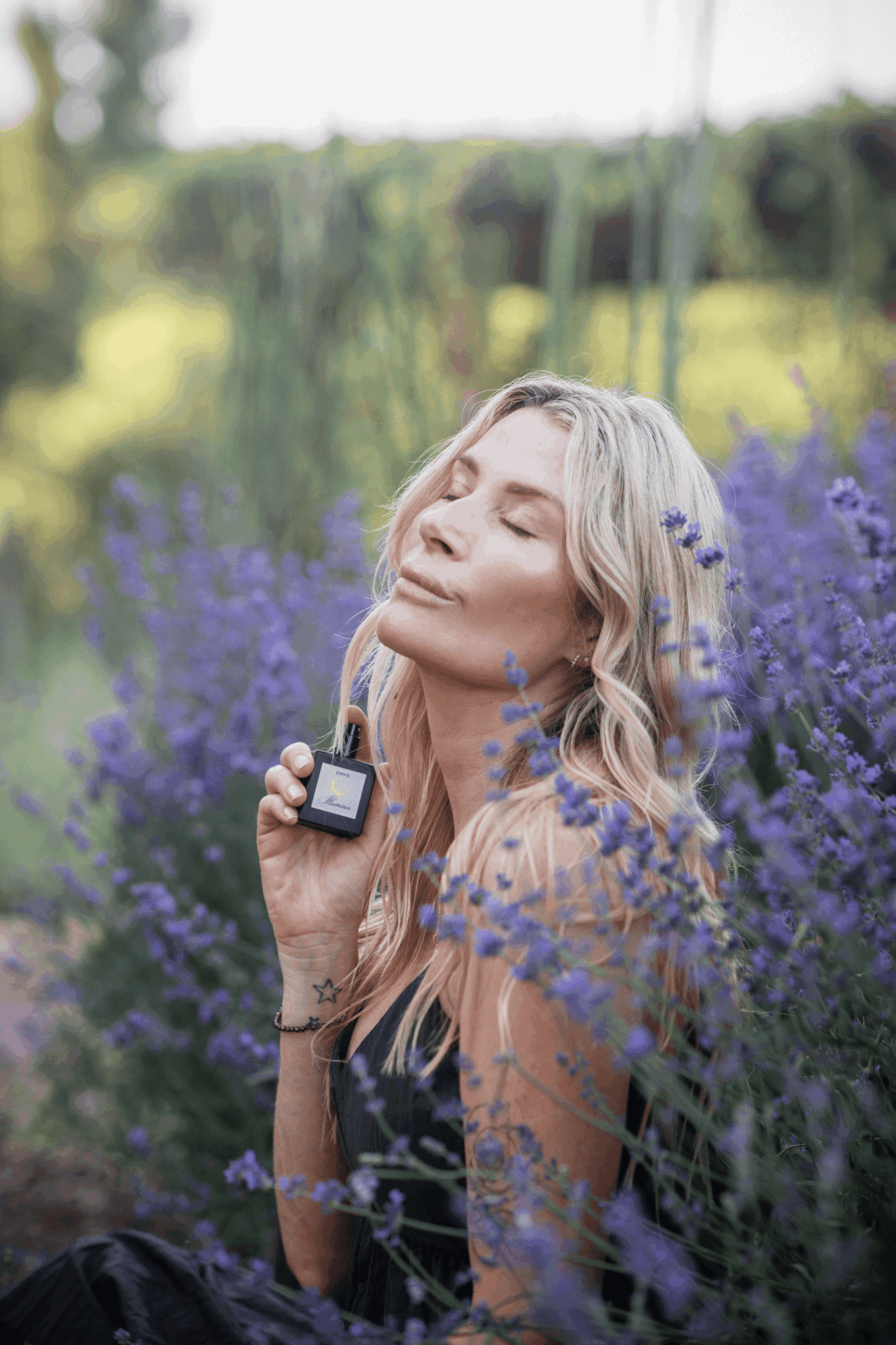 Young woman with blonde hair in a lavender field, holding a small makeup mirror, eyes closed, peaceful expression.