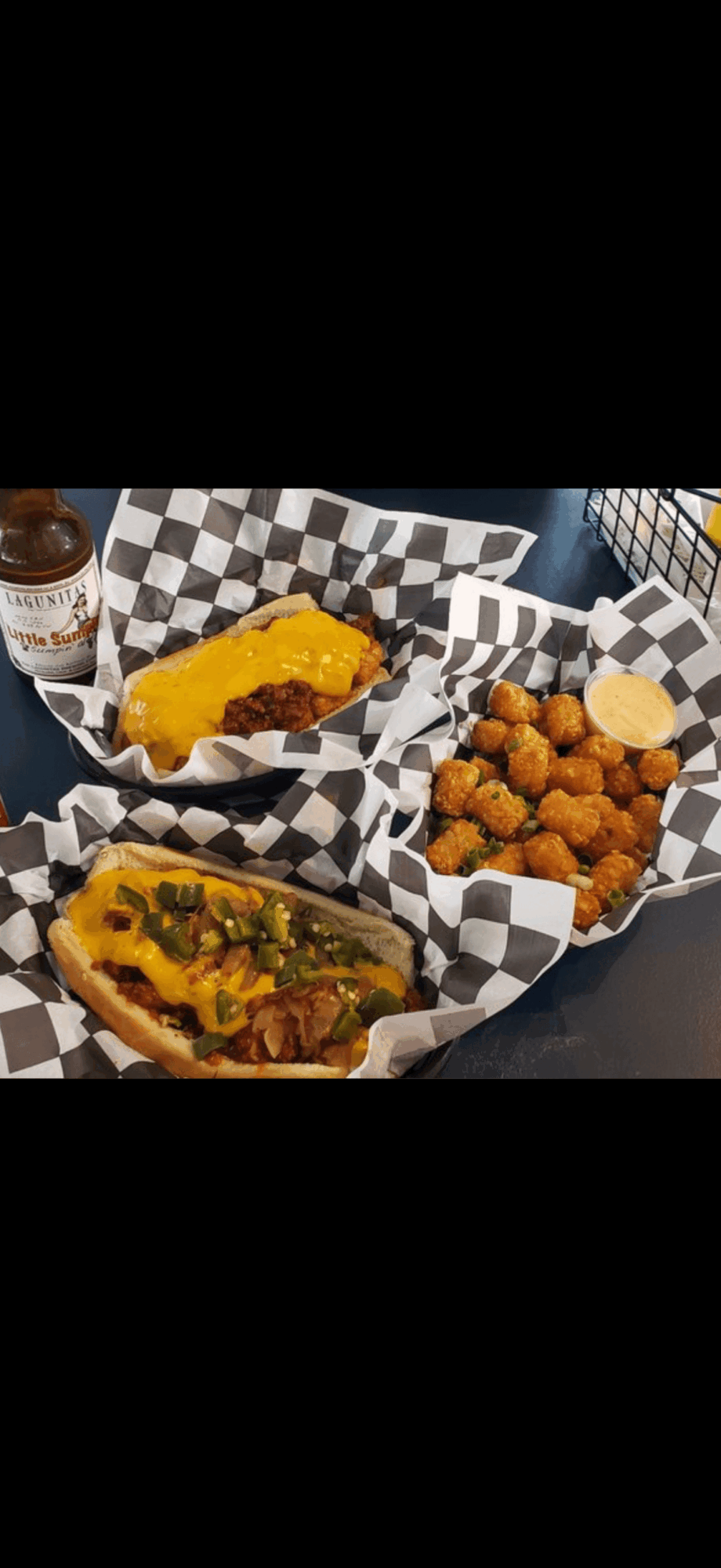 Two cheeseburgers with toppings and a side of fried chicken bites in checkered baskets, with a drink in the background.