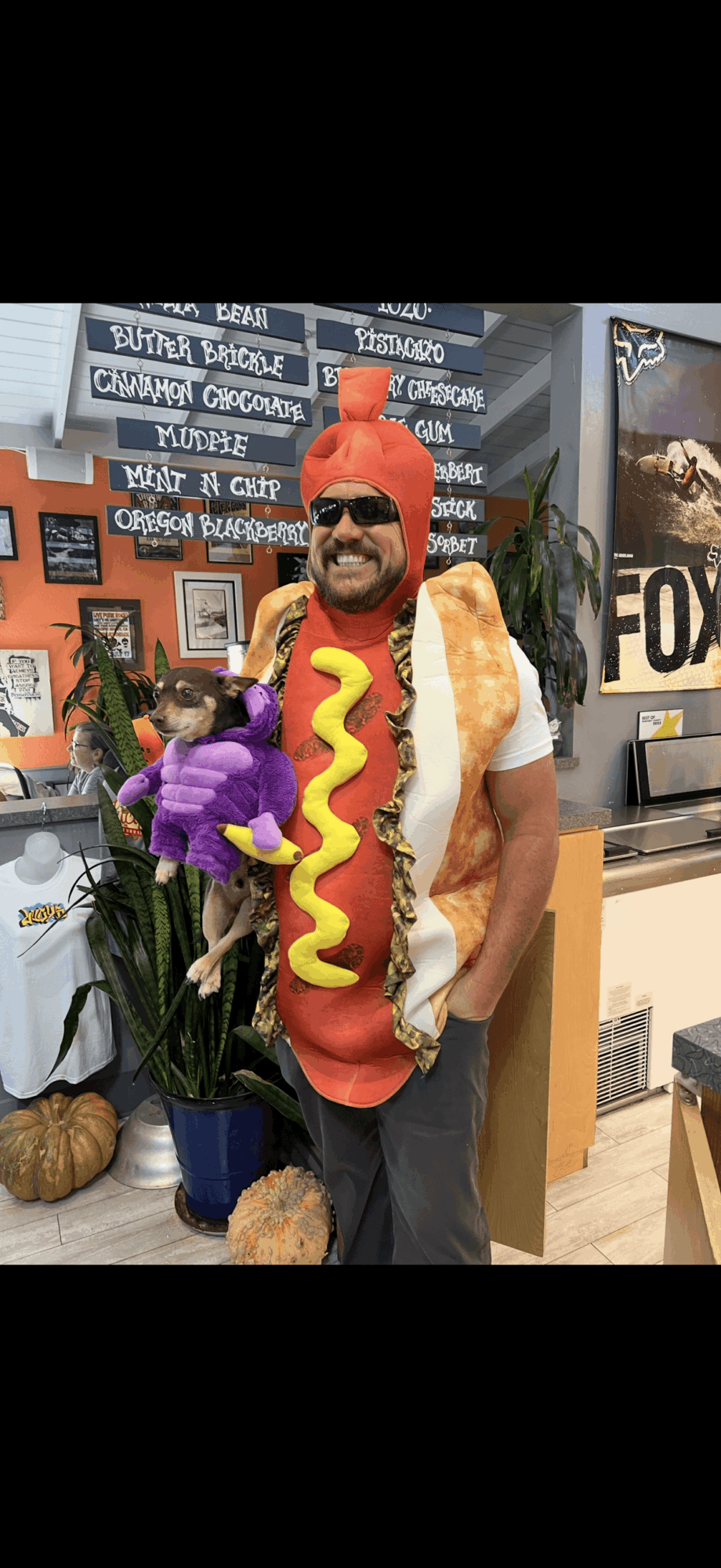 Person dressed in hot dog costume with orange hat, sunglasses, and purple flowers in a store setting.