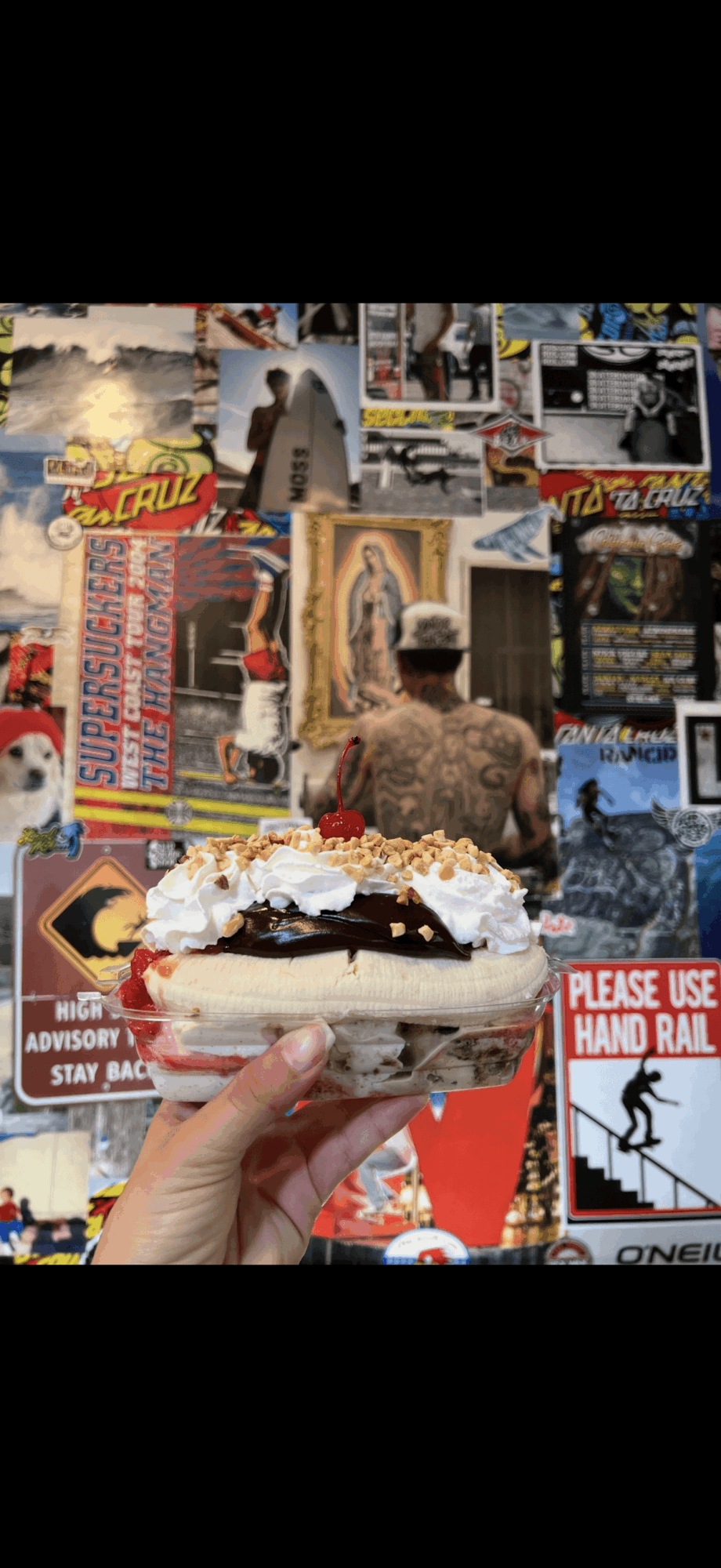 Hand holding a dessert with whipped cream, chocolate, and fruit in front of a colorful collage of posters and images.