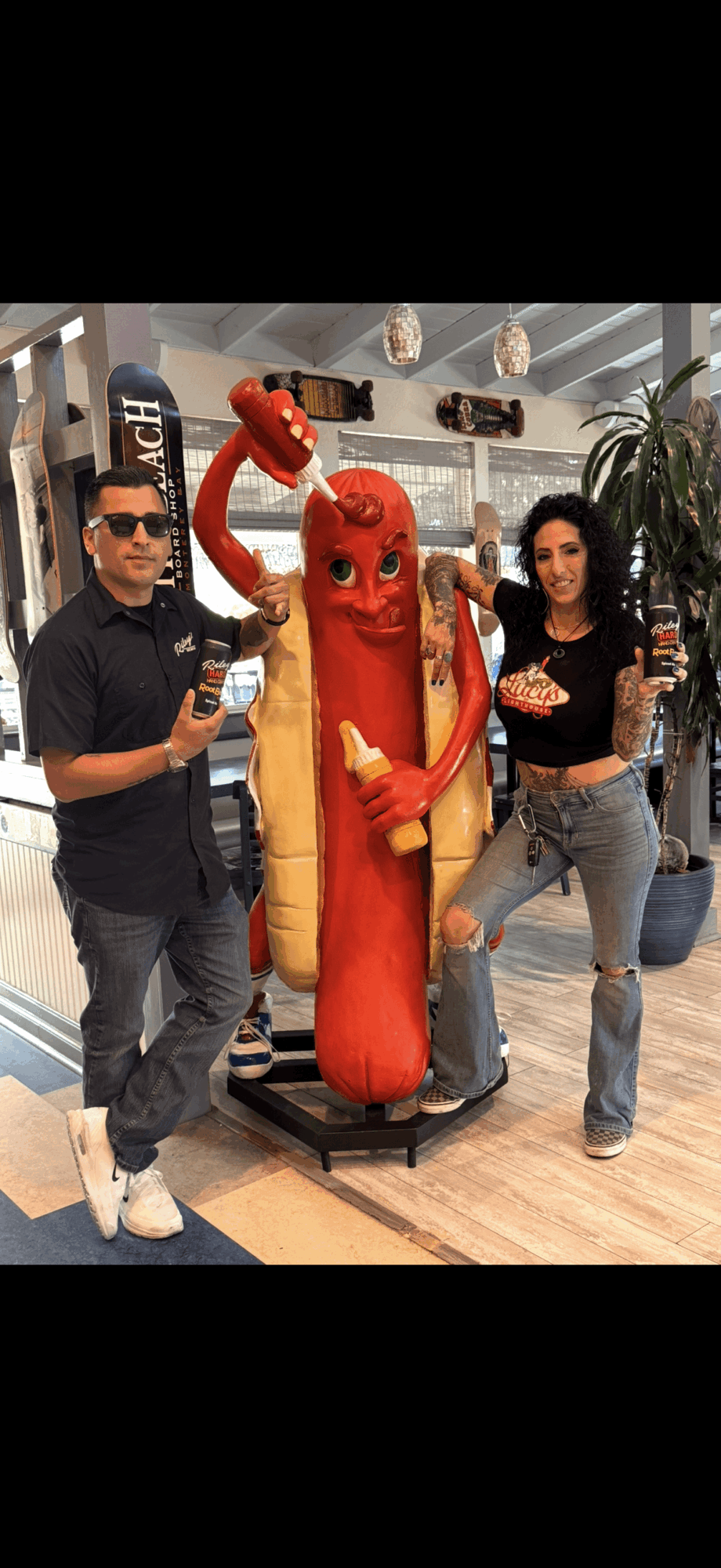 Two people stand next to a hot dog mascot in a shopping mall, holding drinks and posing for the photo.