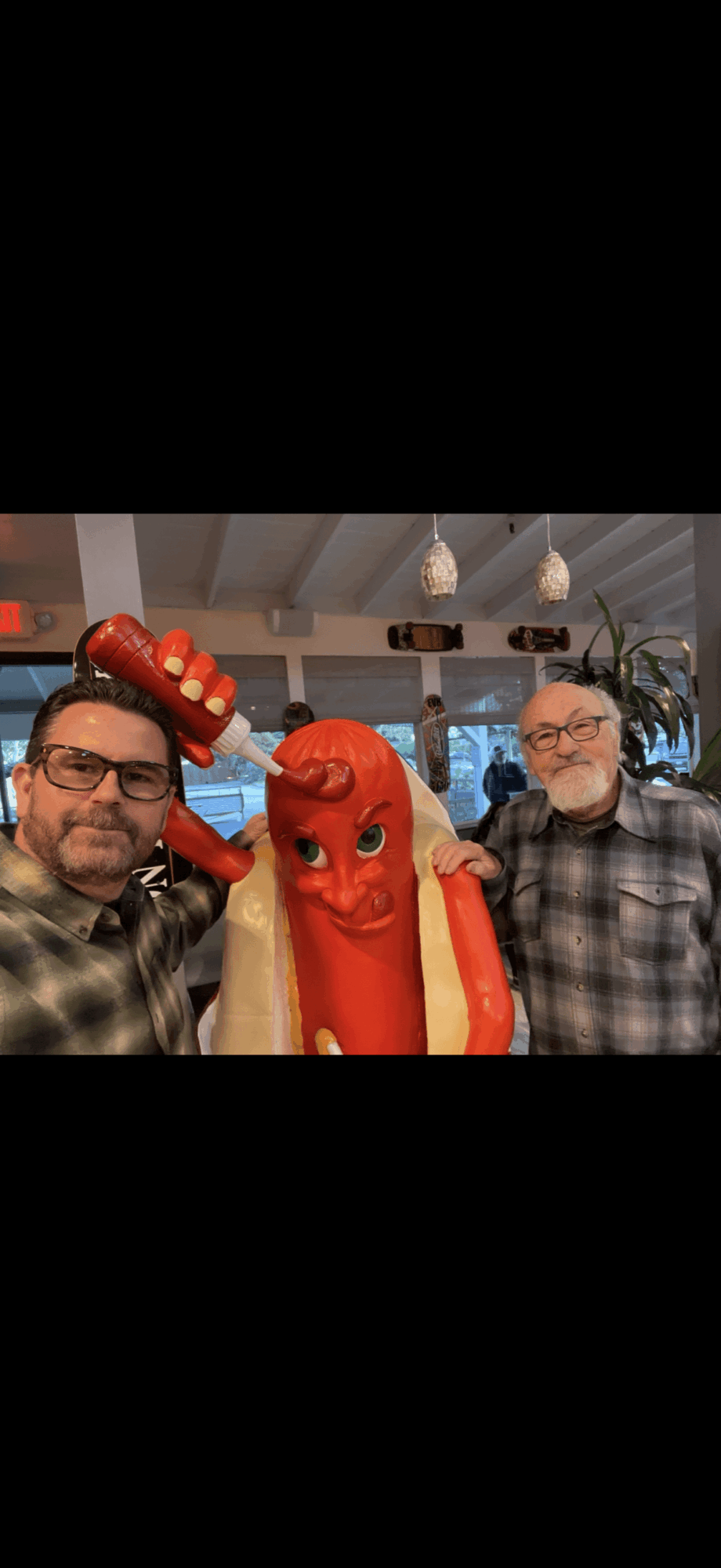 Three people posing with a red inflatable character and a person in a red hat, indoors with hanging lights and plants.