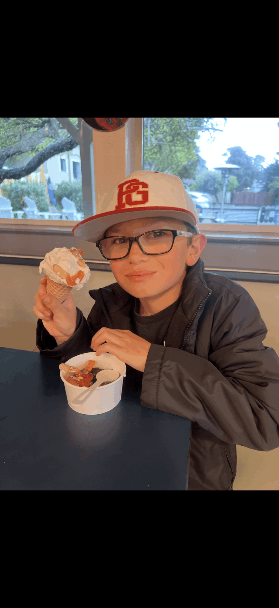Child wearing glasses and a baseball cap, holding ice cream in a cup, sitting at a table indoors with a window view.