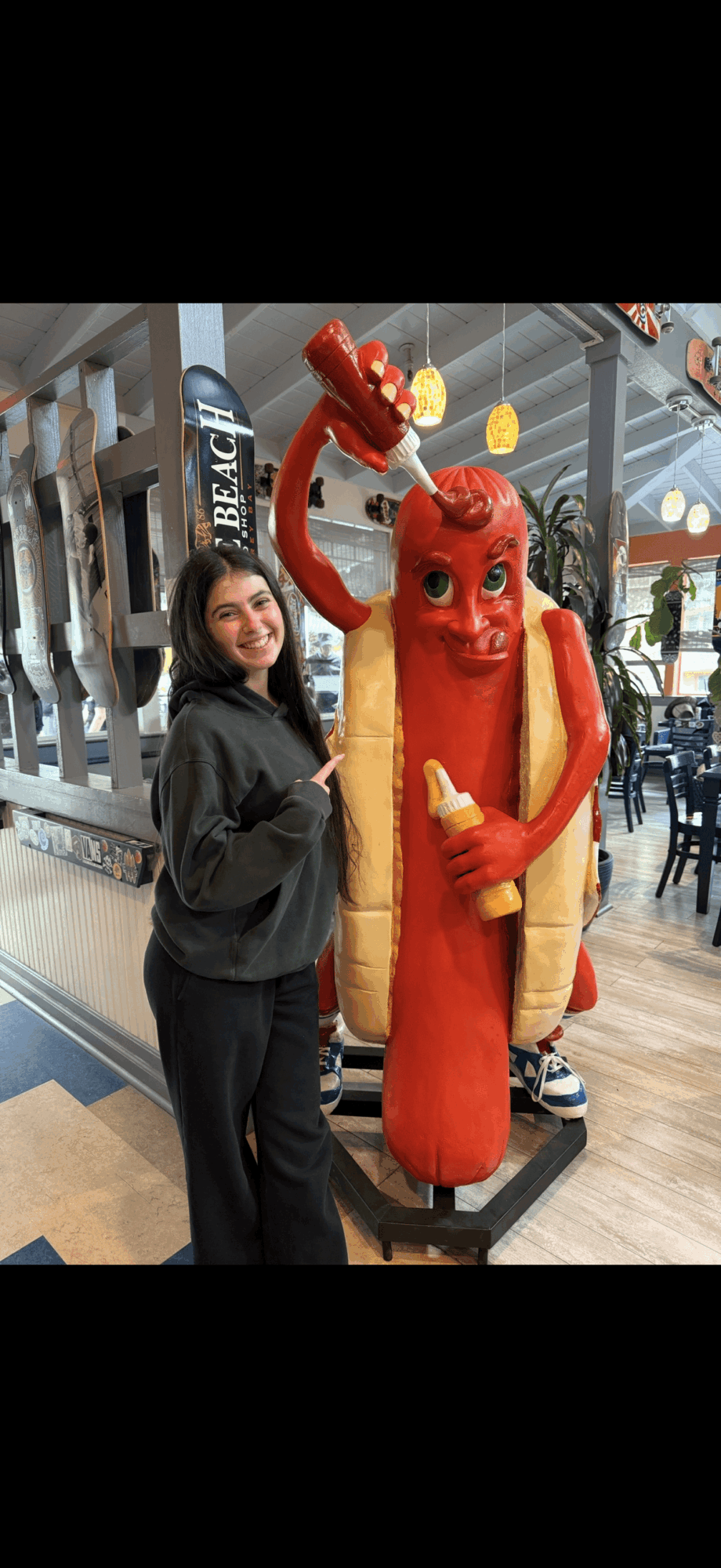 Person standing next to a large red hot dog statue with a smiling face, holding a yellow mustard bottle, inside a shopping mall.