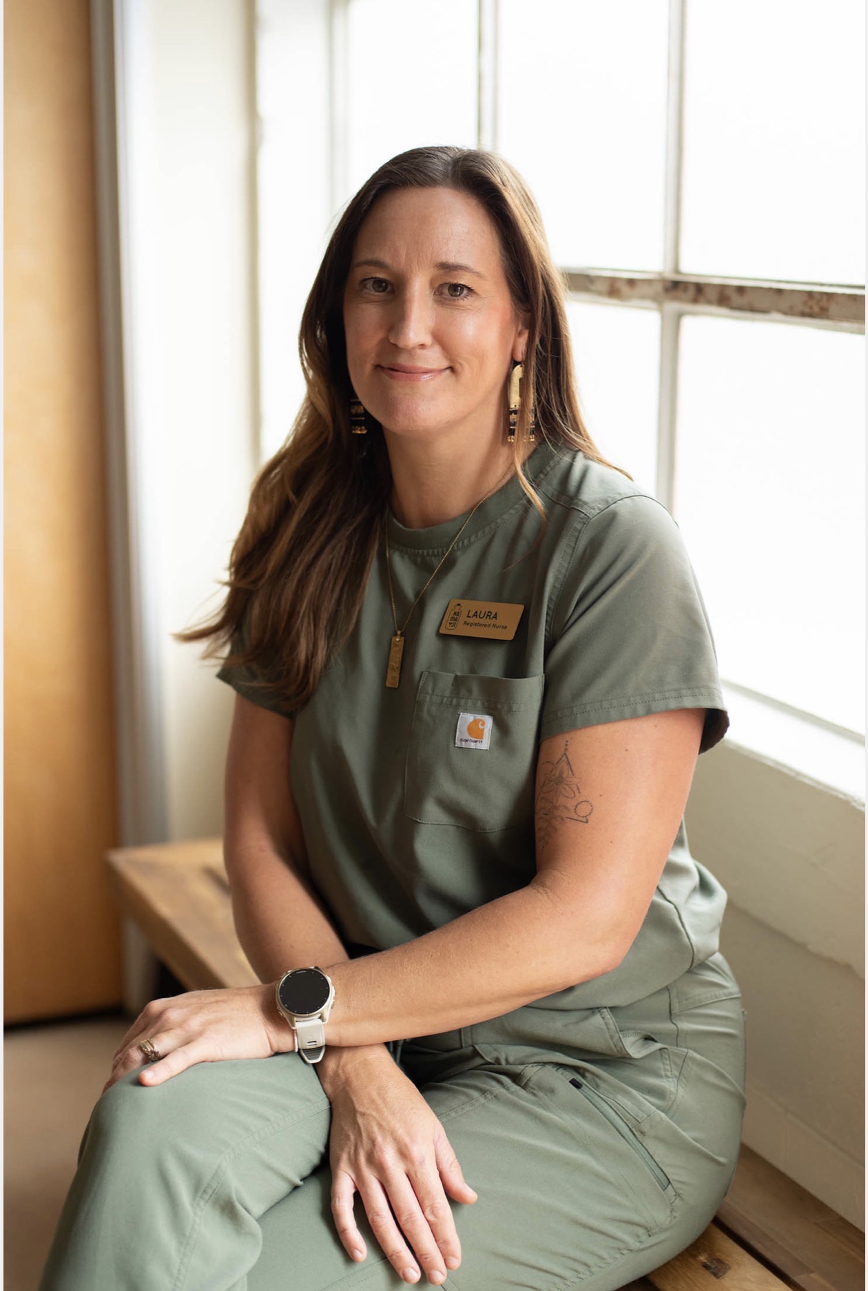 Woman with long brown hair wearing green scrubs, sitting by a window, smiling, with a name tag and smartwatch.
