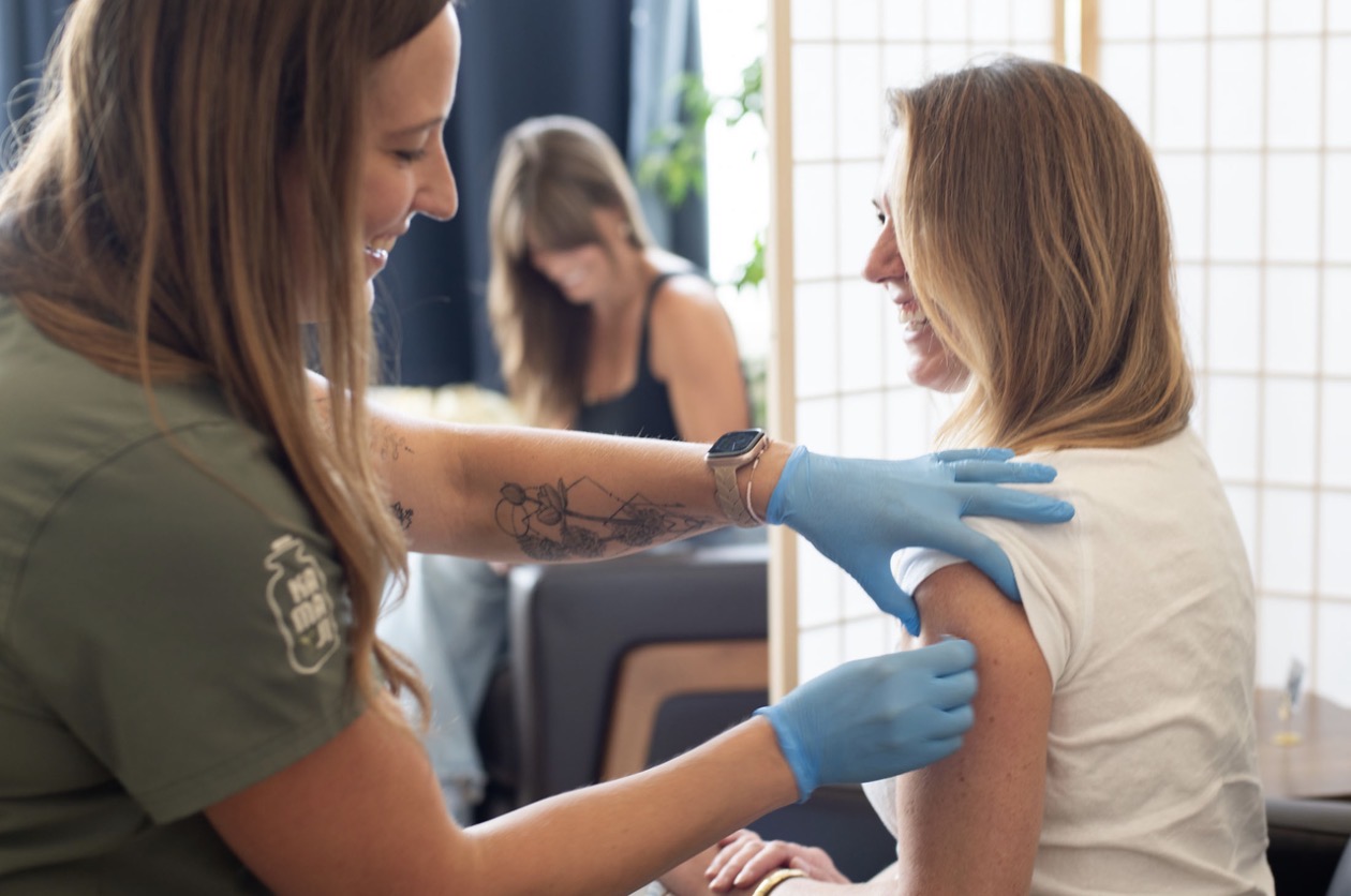 A healthcare worker administers a vaccine to a woman, with another person in the background. The setting appears clinical.