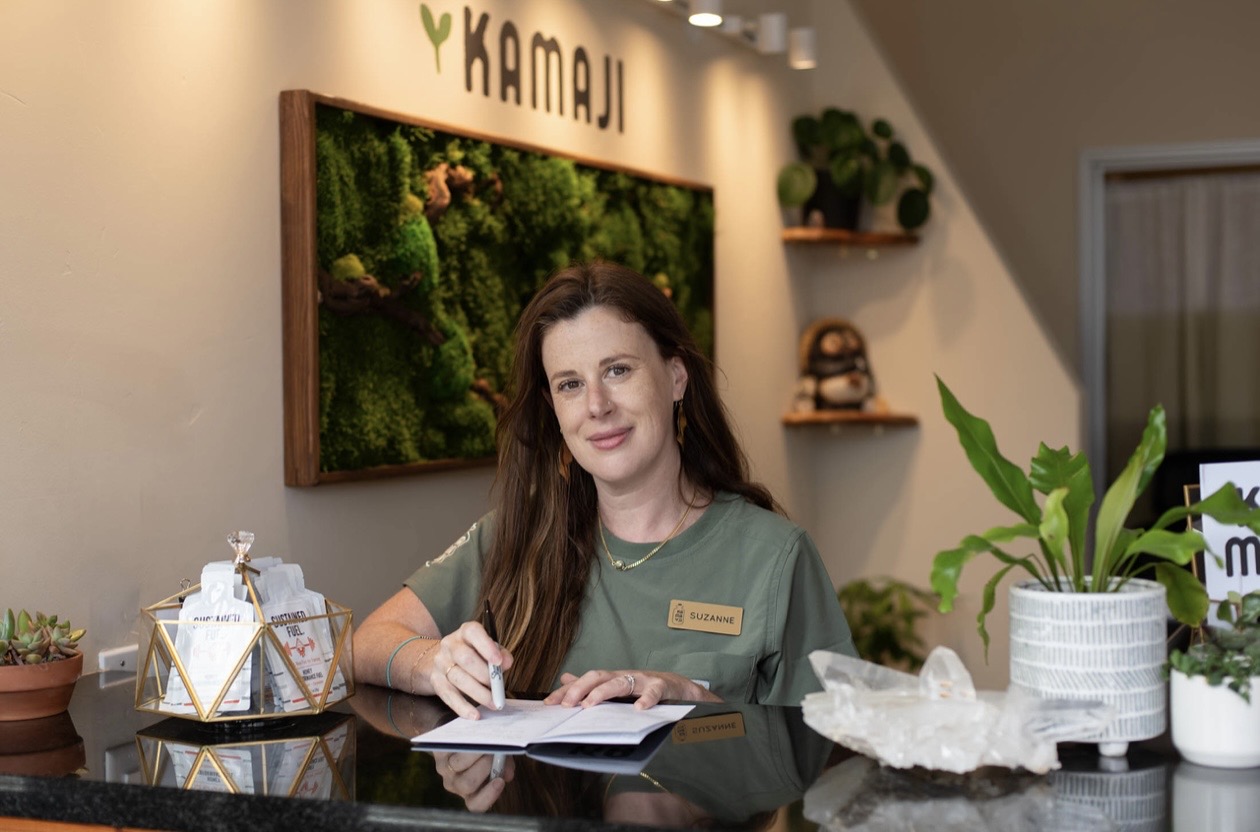 Woman with long brown hair sitting at a desk, smiling, with plants and decorative items in the background.