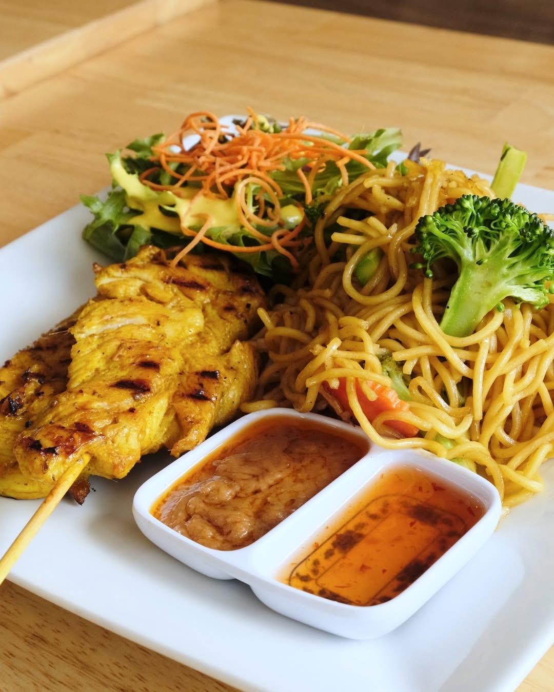 Grilled chicken skewer, noodles with broccoli, and salad with shredded carrots on a white plate, with two dipping sauces.