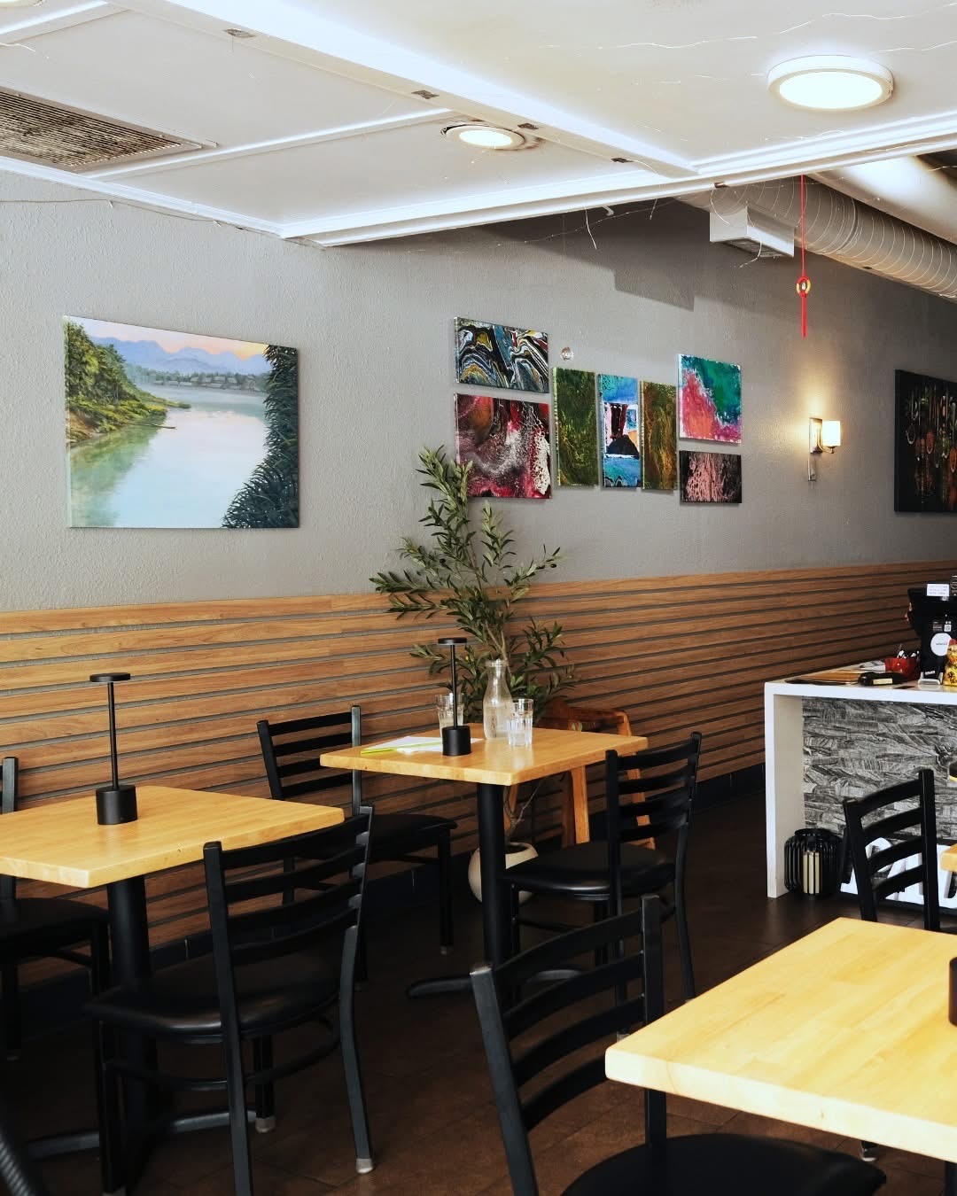 Interior of a cafe with wooden tables, black chairs, and paintings on the wall, including a river scene and colorful abstract art.