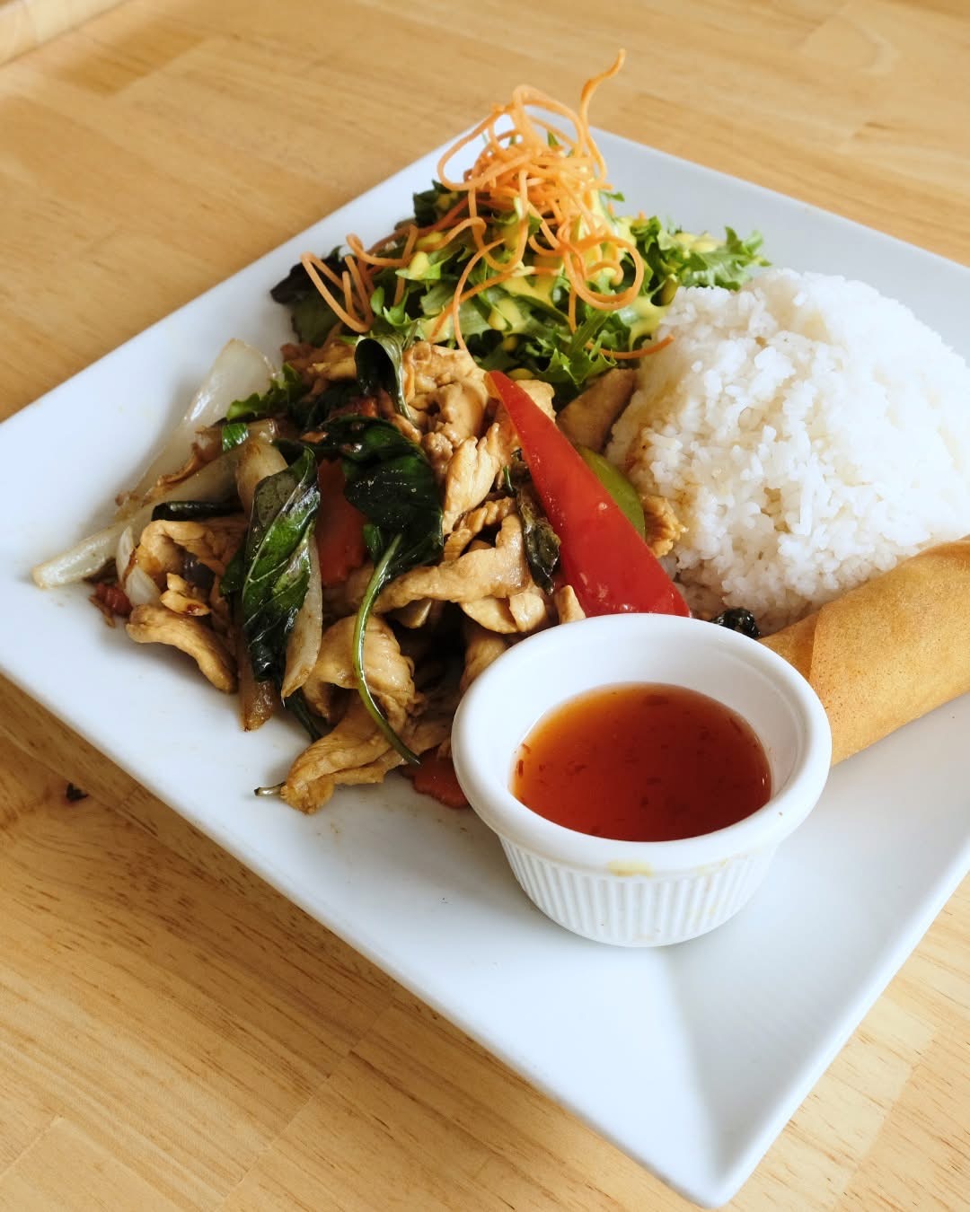 Plate with rice, stir-fried vegetables and meat, salad, and dipping sauce on a white square dish.