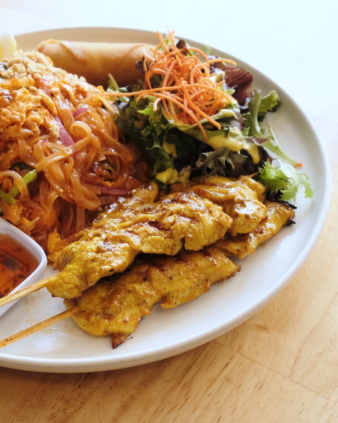 Plate with grilled chicken skewers, salad with shredded carrots, and noodles, on a wooden table.