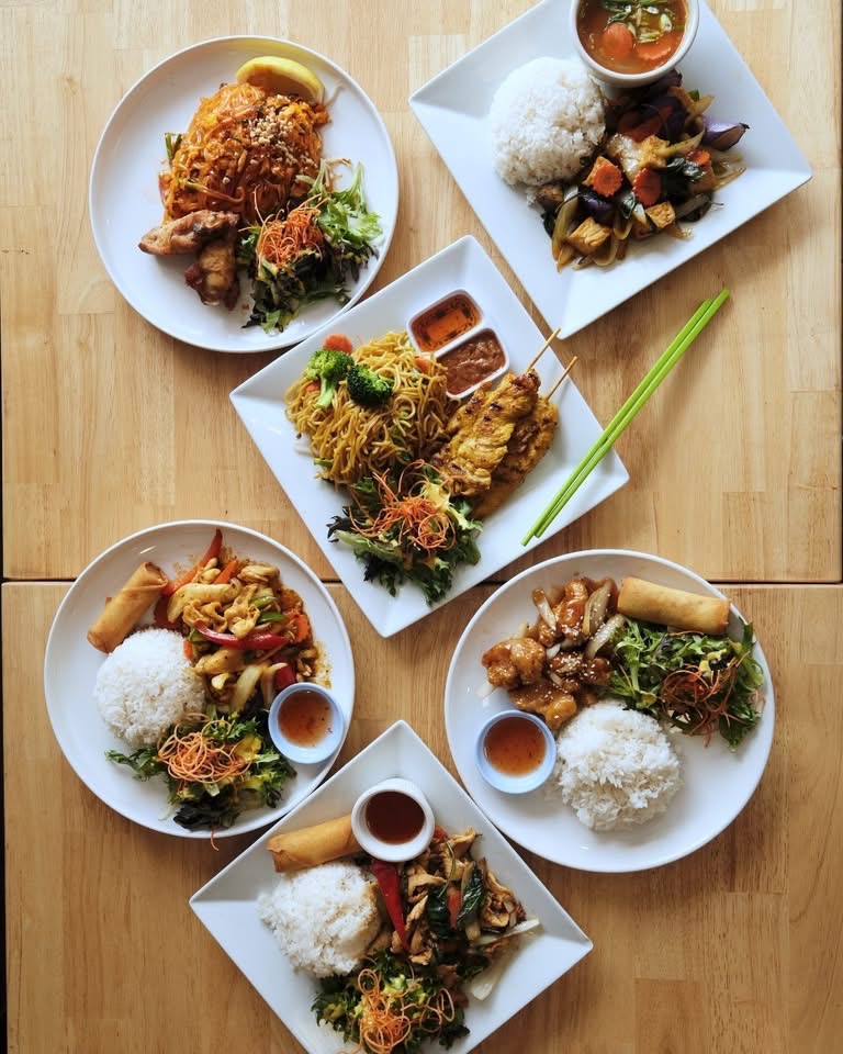 Five plates of Asian dishes with rice, noodles, vegetables, and sauces on a wooden table.