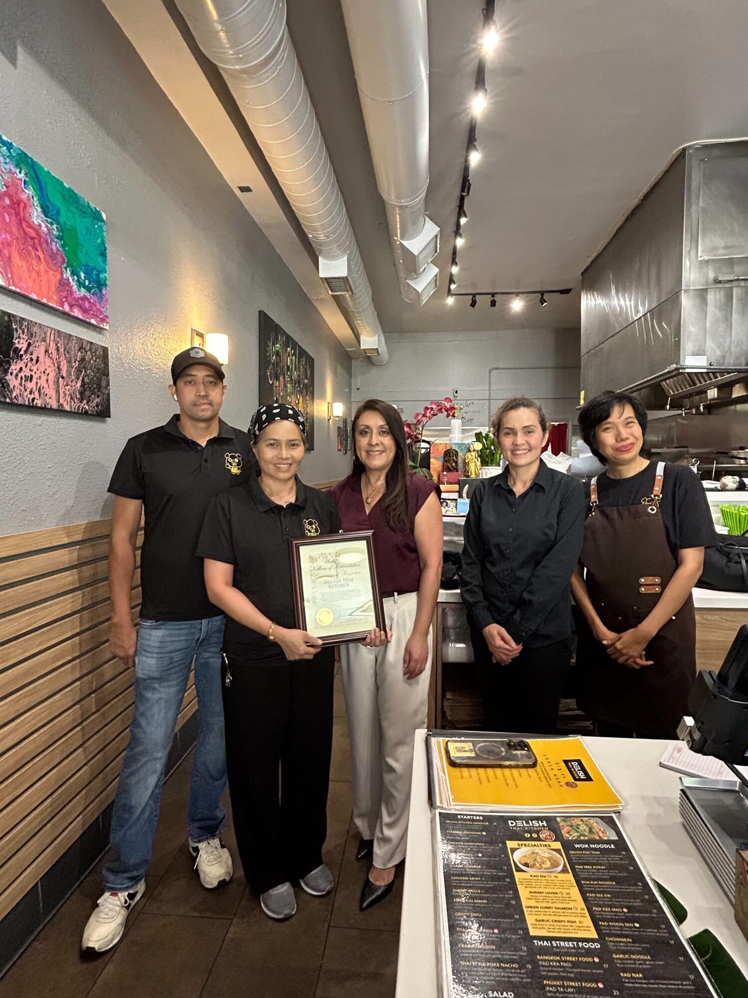 Four people standing inside a restaurant, one holding a certificate, with tables and artwork on the wall behind them.