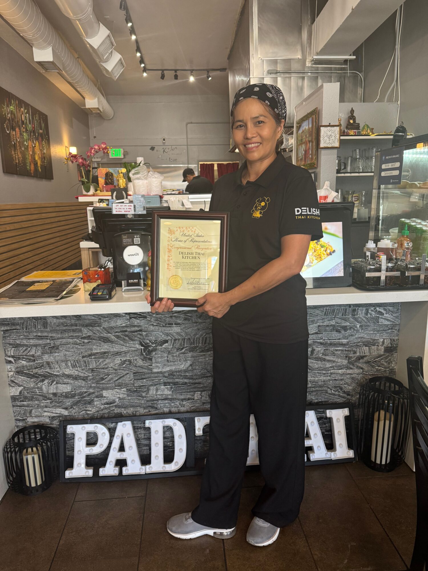 Person in black uniform holding a framed certificate inside a cafe or restaurant, standing on a mat with the word 'PAID'.
