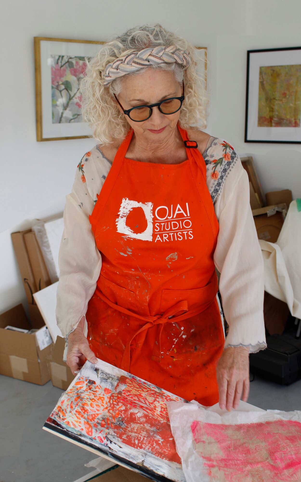Woman with curly hair and glasses wearing a red apron, looking at artwork on table, in a room with framed pictures.