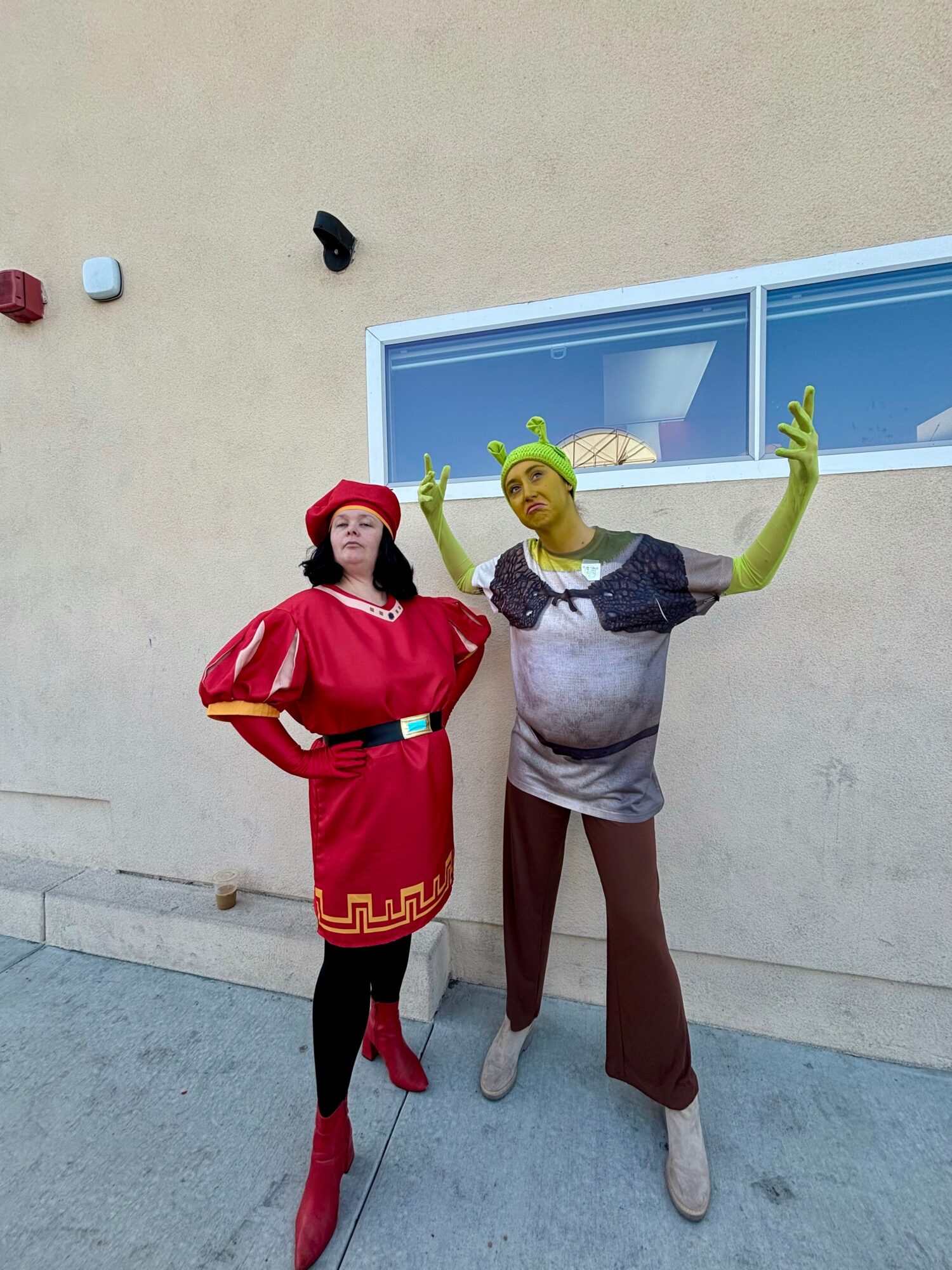 Two people in colorful costumes stand outside a beige wall with a window, one with green skin and the other in red.