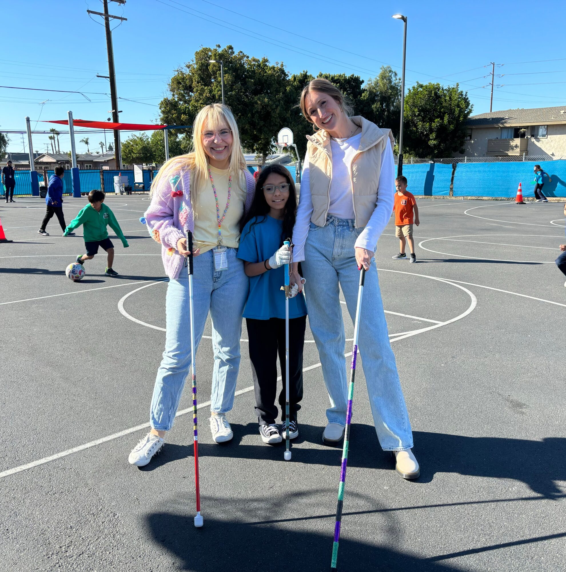 Three people standing on a basketball court, two women and a girl, smiling, with children playing in the background.
