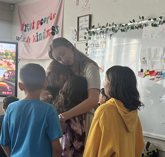 Teacher interacts with three children in a classroom, with a colorful banner and whiteboard in the background.
