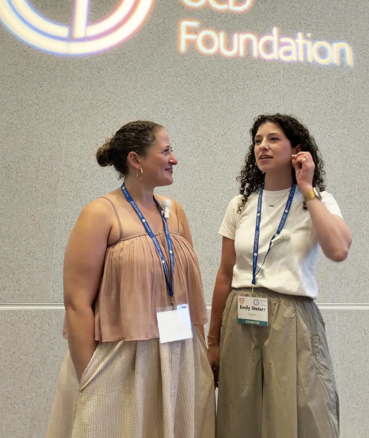 Two women standing and talking at an event, wearing name tags and lanyards, with a logo and text in the background.