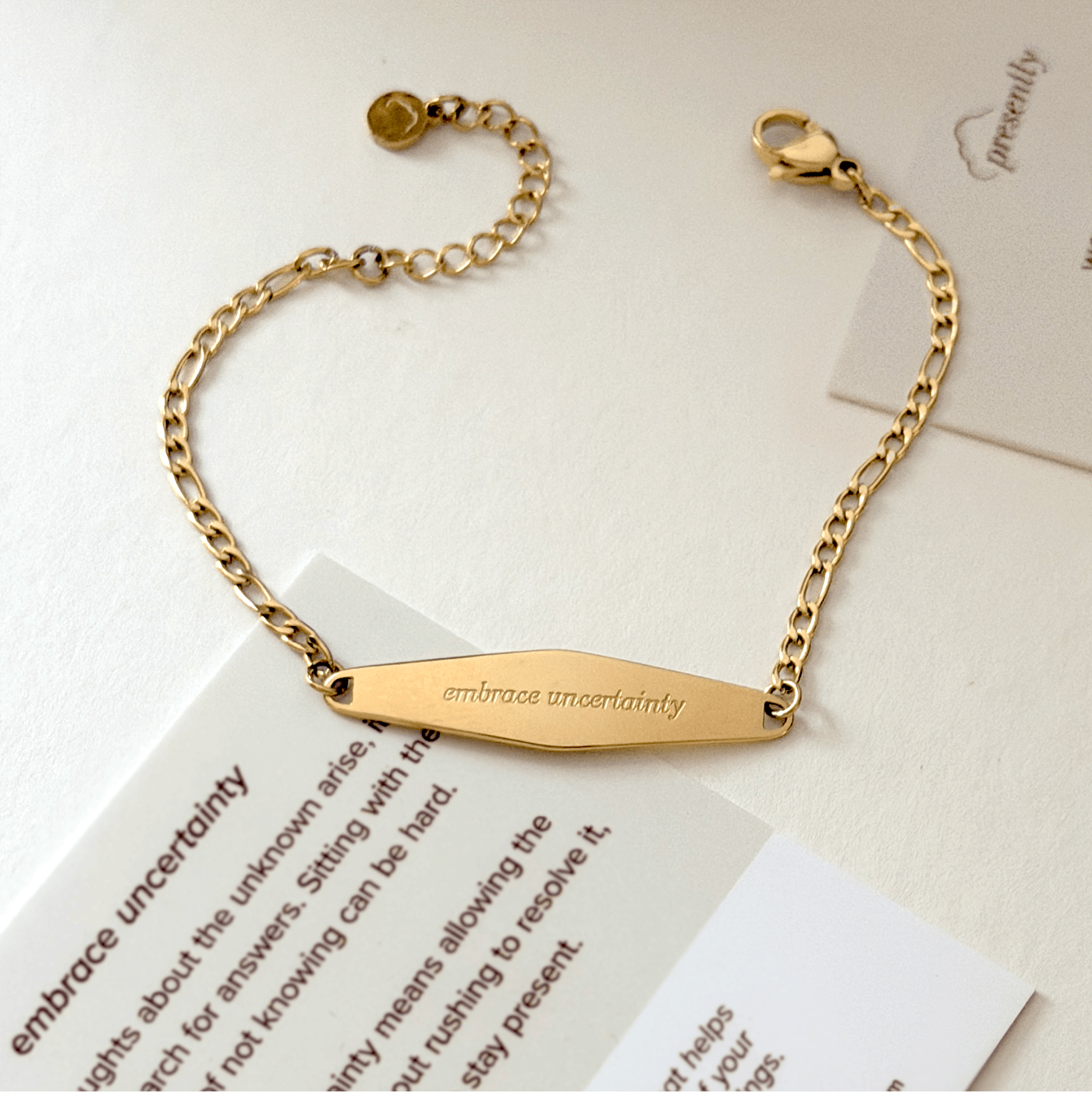 Gold bracelet with a bar engraved 'embrace uncertainty' on a white surface with papers nearby.