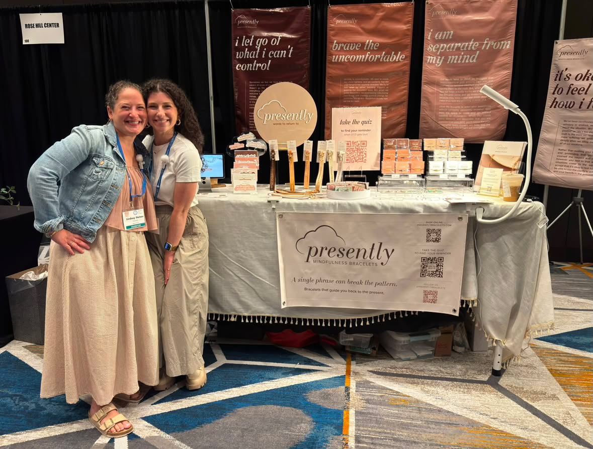 Two women stand smiling in front of a booth with skincare products and a banner reading 'presently'.