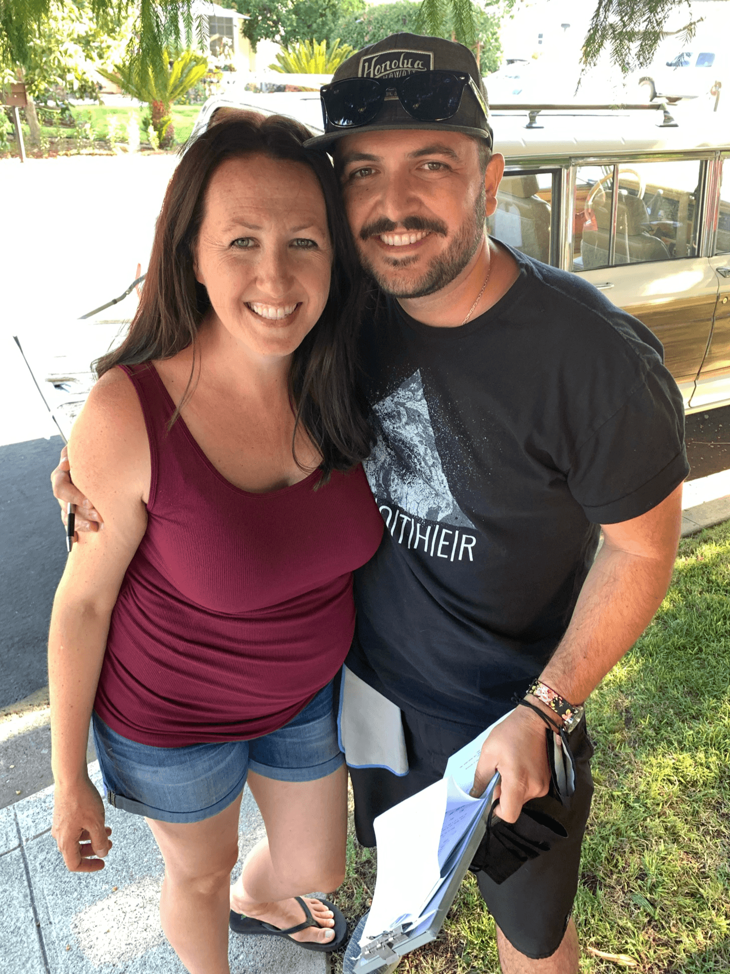 A smiling woman and man stand outdoors, close together, with trees and a vehicle in the background.