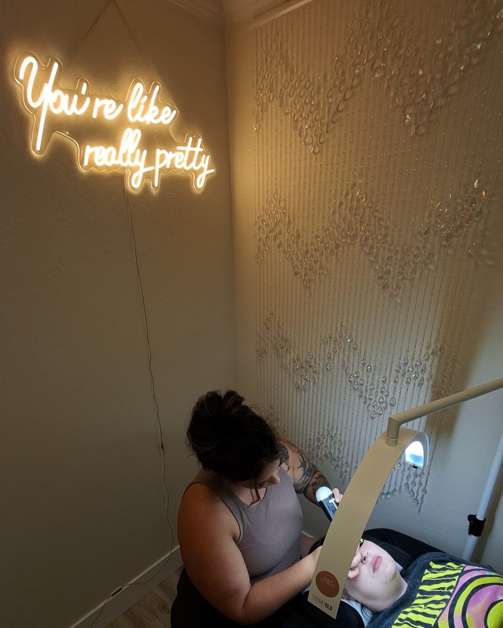 Neon sign reads 'You're like really pretty' on a wall, with a woman sitting on stairs looking at her phone.