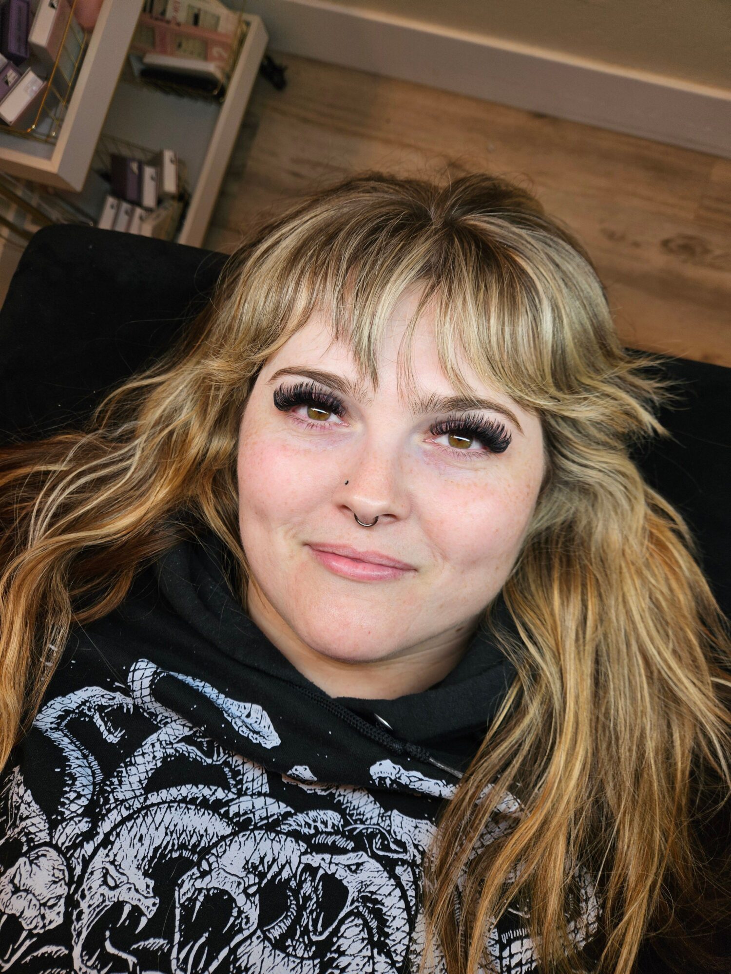Woman with long, wavy blonde hair lying on a black surface, smiling, with a patterned top and a wooden floor background.